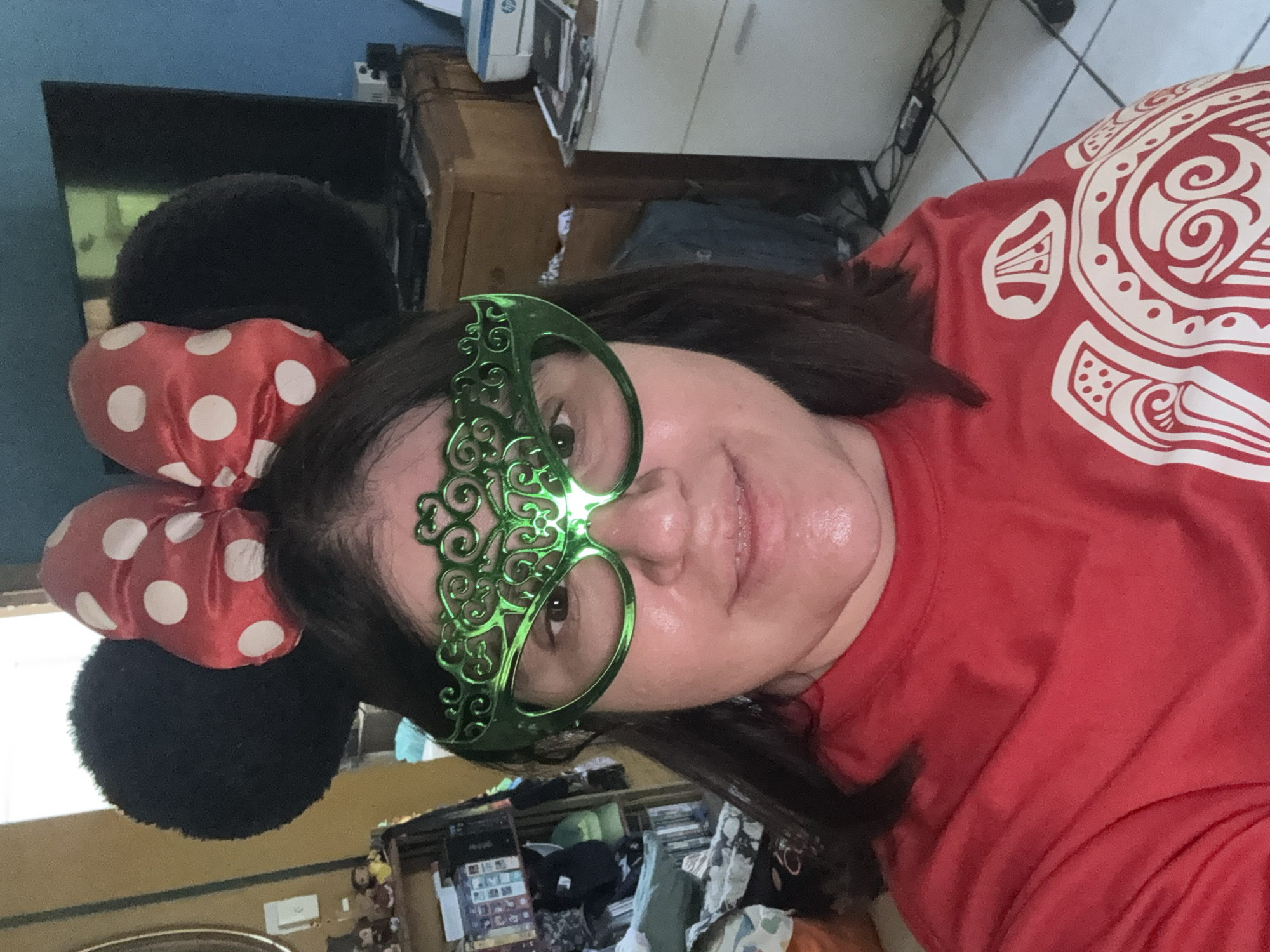 A close-up, rotated photo captures a woman indoors, looking directly at the camera with a gentle, pleasant expression. She is adorned in a playful costume, featuring black Minnie Mouse ears topped with a red and white polka-dotted bow, and a bright green, intricately designed masquerade-style mask that covers her eyes. The mask has a light source at the bridge, casting a subtle glow on her face. Her dark hair is visible around her head and shoulders. She wears a vibrant red t-shirt embellished with a large white graphic design, which appears to be a stylized tribal or Polynesian pattern.
The background suggests a casual, lived-in home environment in Xochitepec, Mexico. To the left, a blue wall is visible, behind which is a dark screen or monitor. To her right, a wooden dresser or cabinet holds various household items, including a white and blue HP printer and a scattering of papers, boxes, and other objects, indicating some clutter. The lower background shows a tile floor and more indistinct household items. The overall impression is one of lightheartedness and fun, possibly a selfie taken during a festive moment or simply for amusement. The only discernible text is the "HP" logo on the printer.