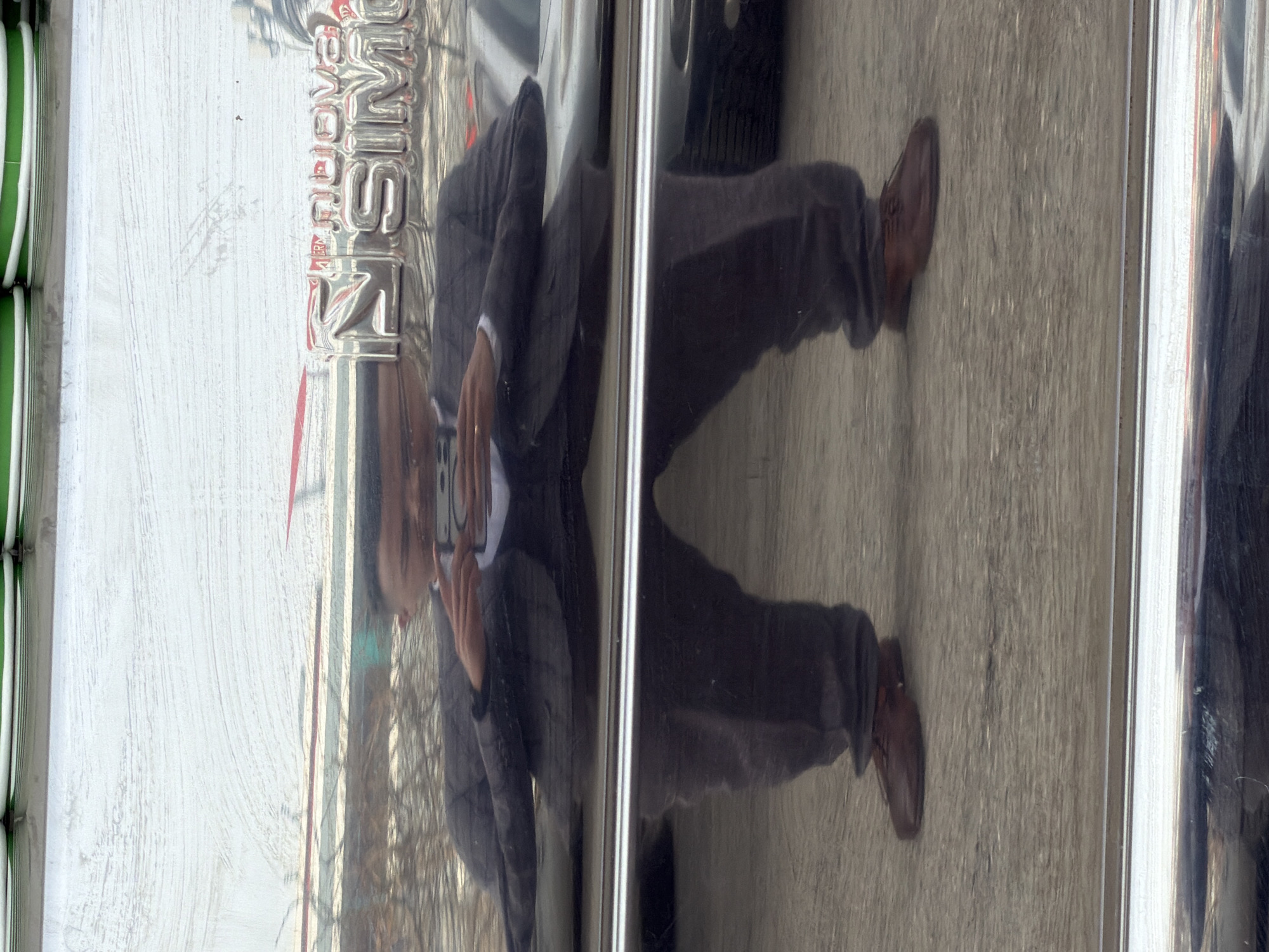Reflection of a man taking a photo with his phone on a shiny surface with the text "PERM CITY" on the upper left. The man is wearing a suit jacket, dress pants, and brown dress shoes. The background appears to be a paved sidewalk or street. The image was taken in Perm, Russia.
