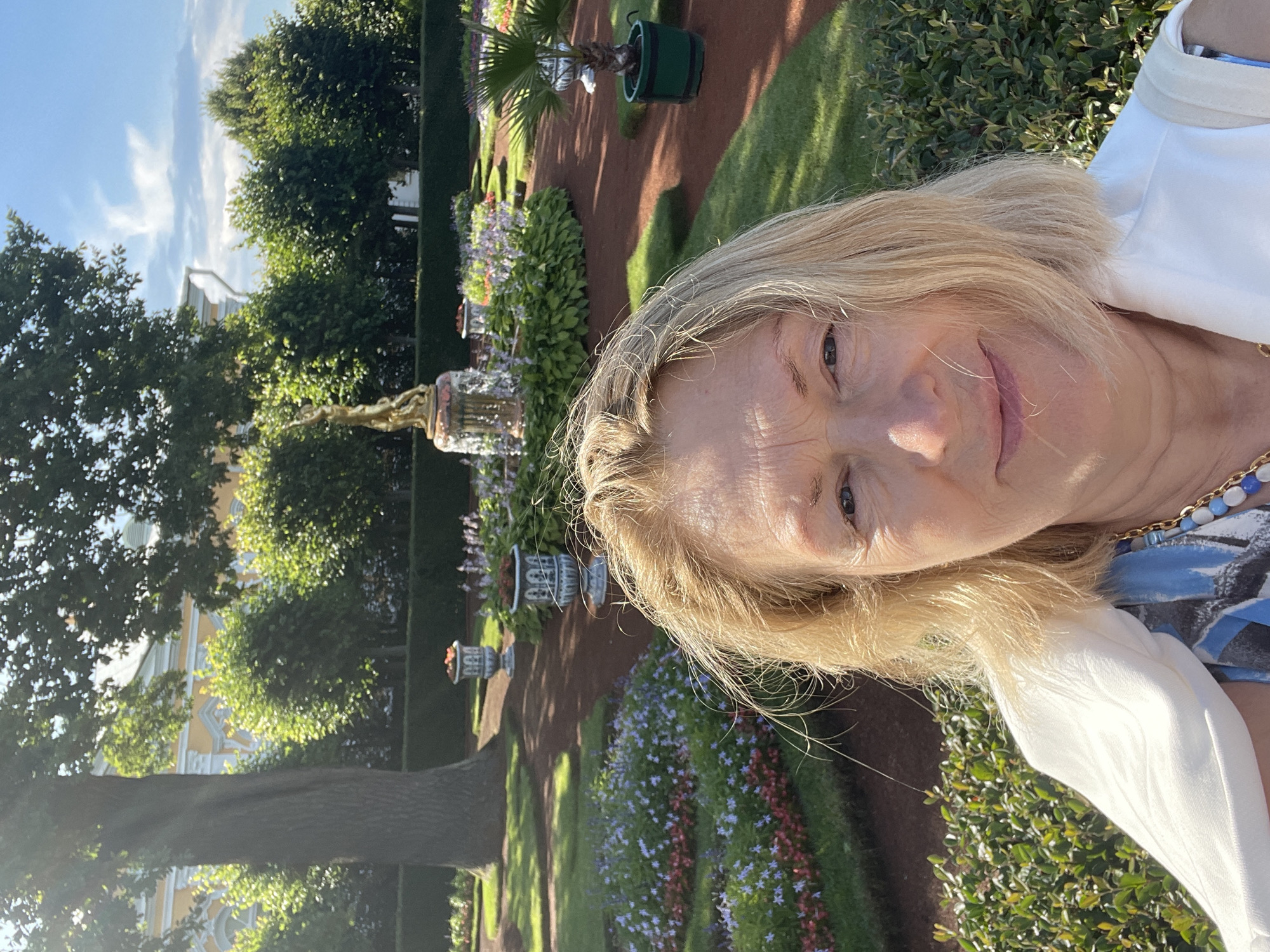 The image is a selfie taken outdoors in the city of Peterhof, Russia, likely within the Peterhof Palace complex. The foreground is dominated by the face of a woman with blonde hair and fair skin, looking towards the camera. She is wearing a white garment draped over her shoulders and a blue and white patterned top with a gold necklace. The background features a meticulously maintained formal garden.
The garden is characterized by manicured green lawns, geometric flower beds with red and purple flowers, and well-trimmed hedges. Several ornate urns are visible, some containing flowers. In the mid-ground, a golden statue stands atop a fountain, surrounded by lush greenery and more flowers. Further in the background, a large tree trunk is prominent on the left, and a yellow building with white architectural details, possibly part of the palace, is visible through the foliage on the left side of the frame. The sky is blue with scattered white clouds. The lighting suggests it is a sunny day.