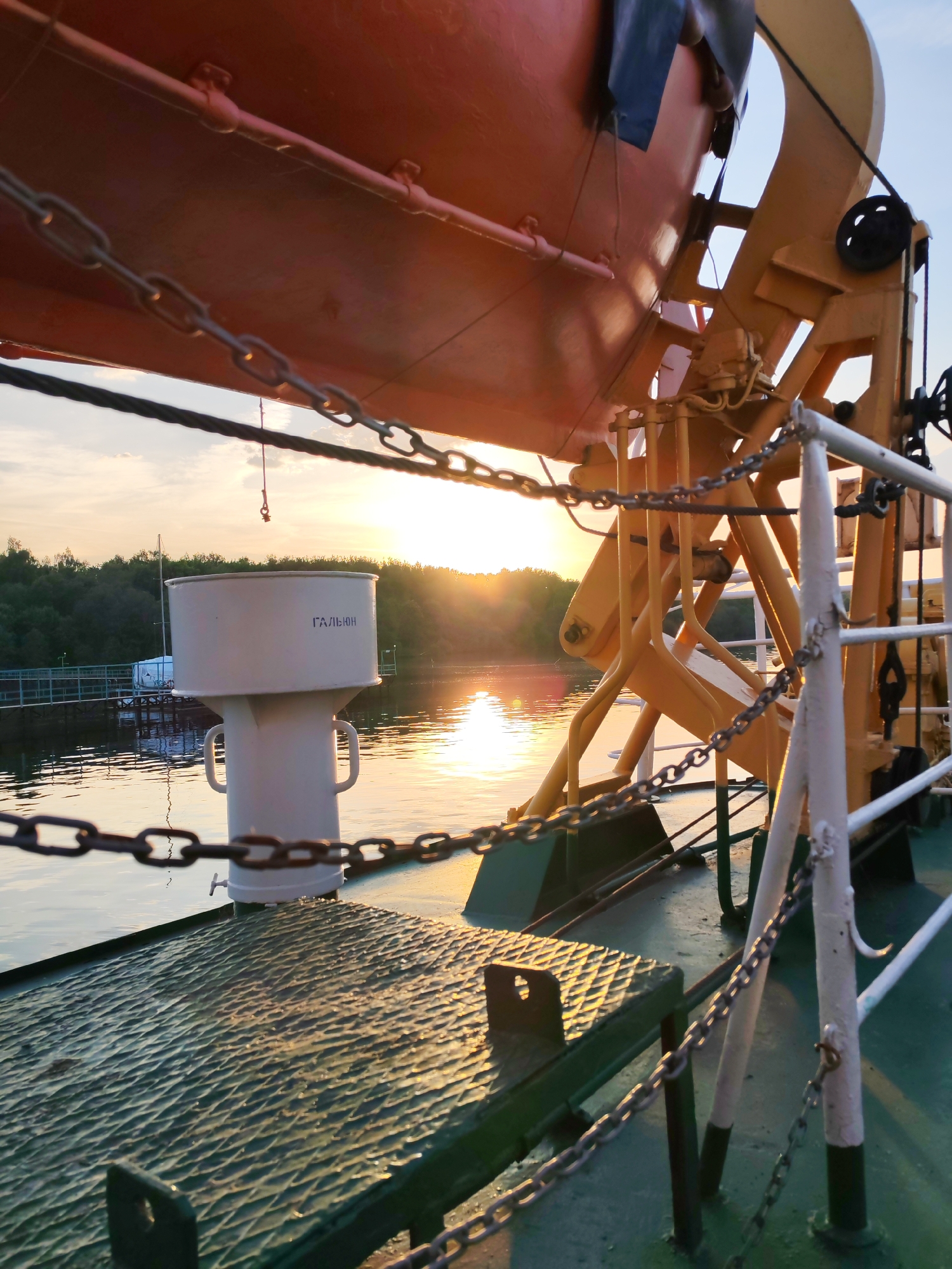 Aboard a vessel in Ermolino, Russia, the scene captures a sunlit view across a body of water toward a wooded shoreline. The ship's deck is visible in the foreground, featuring a patterned green surface. A white structure labeled "ГАЛЬЮН" in Cyrillic is affixed to the deck. The ship also contains orange safety equipment suspended by yellow supports, with chains forming part of the safety barrier. The low sun reflects on the water's surface, indicating either sunrise or sunset.