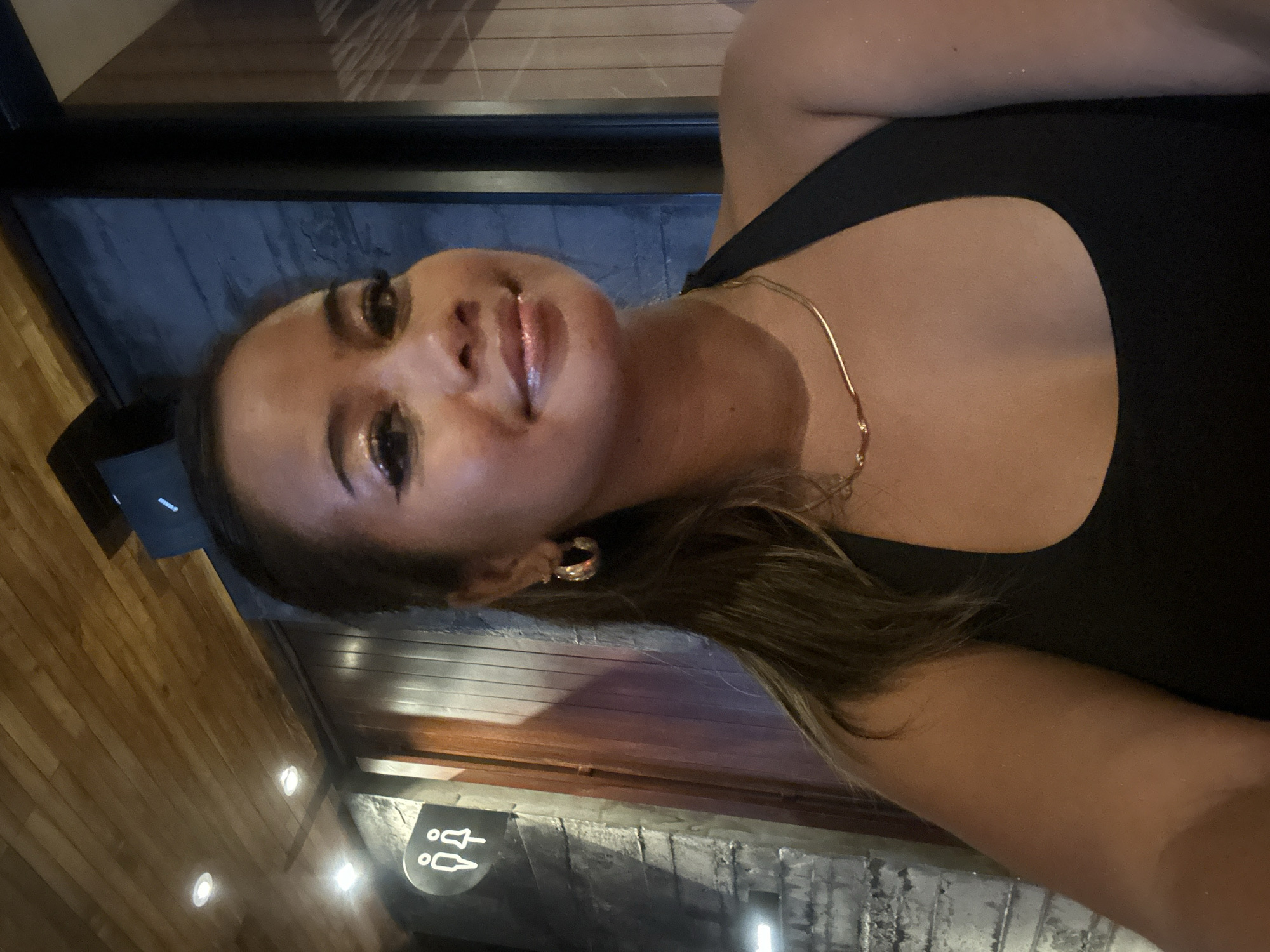 A medium close-up, tilted selfie captures a young woman within what appears to be a dimly lit indoor establishment. She looks directly at the camera with a gentle smile, her head playfully tilted. Her face features clear skin, defined eye makeup with a hint of shimmer, and glossy lips. Her dark brown hair falls over her shoulder, complementing her black, sleeveless top or dress. She wears a delicate gold chain necklace and a noticeable gold hoop earring on her visible ear.
The background shows a mix of textures: warm wooden paneling on the left and ceiling, and a darker, possibly stone or textured block wall on the right. Recessed spotlights provide ambient illumination, suggesting an evening or nighttime setting. In the bottom right corner, a sign featuring stylized male and female figures, commonly used for restrooms, is visible. The scene evokes the atmosphere of a modern restaurant or bar, consistent with locations found in North Kuta, Indonesia. The woman's expression conveys a calm and self-assured demeanor.