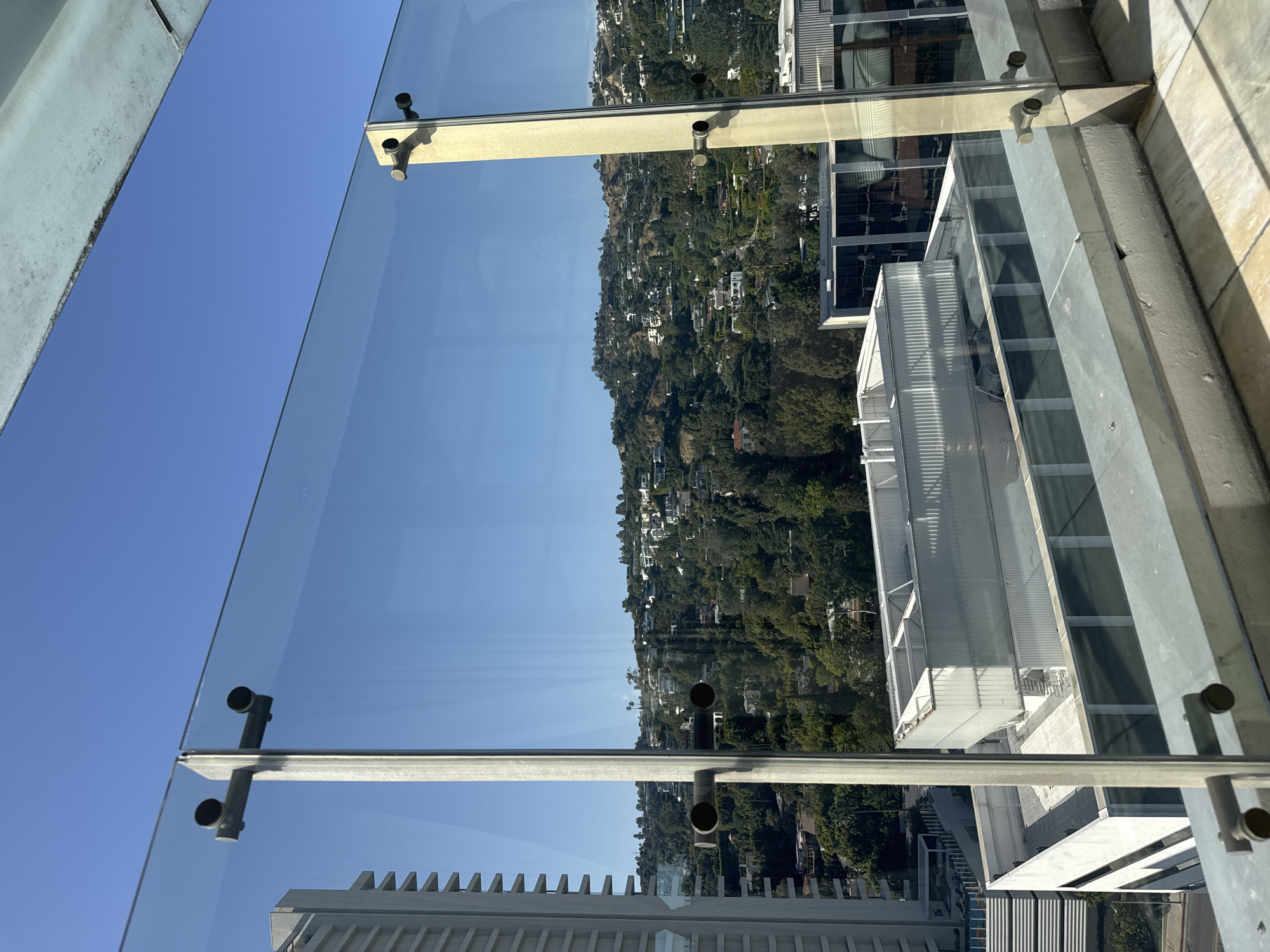 A medium, eye-level, wide-angle shot captures the view from a high-rise in West Hollywood, United States. The foreground features architectural elements: a glass barrier with metallic supports that frame the view. Beyond the barrier, the backdrop reveals a densely wooded hillside, typical of the Hollywood Hills, dotted with houses.
Clear blue sky dominates the upper portion of the frame, reflected in the glass. Below, modern buildings with metallic and glass facades are visible, partially obscured by the angle and perspective.
The scene appears to be captured during daylight hours under clear weather conditions. There are no people or animals visible in the frame. The focus is on the urban landscape blending into the natural terrain of West Hollywood.