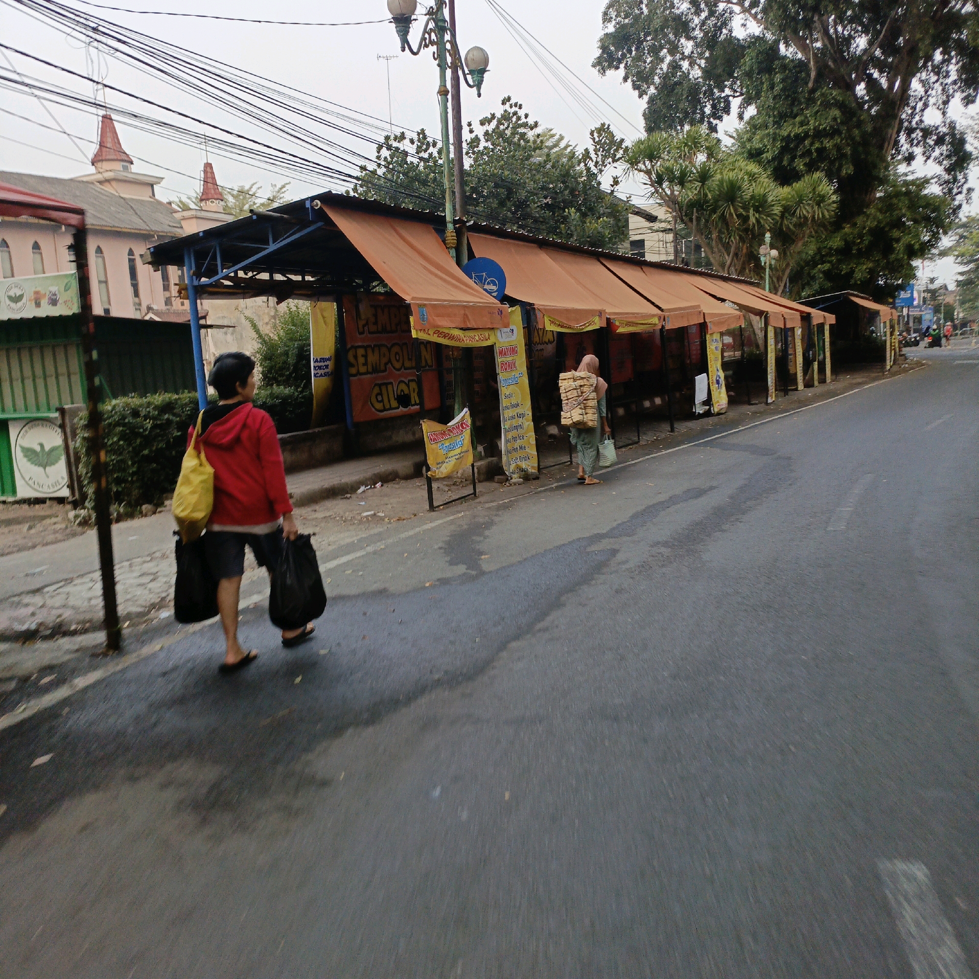 A street scene in Salatiga, Indonesia, features a row of outdoor stalls with orange awnings lining a paved road. A person in a red hoodie and dark shorts walks away from the camera, carrying a yellow bag over their left shoulder and two black plastic bags in their hands. Another person wearing a headscarf and a light-colored outfit is walking away from the camera further down the street, carrying a large stack of items on their back and a light-colored bag in their right hand. The stalls display various signs, including one in yellow that reads "PEMPEK SEMPOLIN CILOR" and another that says "WARUNG ROKOK AGASSI". The environment includes trees, overhead power lines, and a building with distinctive reddish-pink spires in the background. The road appears wet in patches.