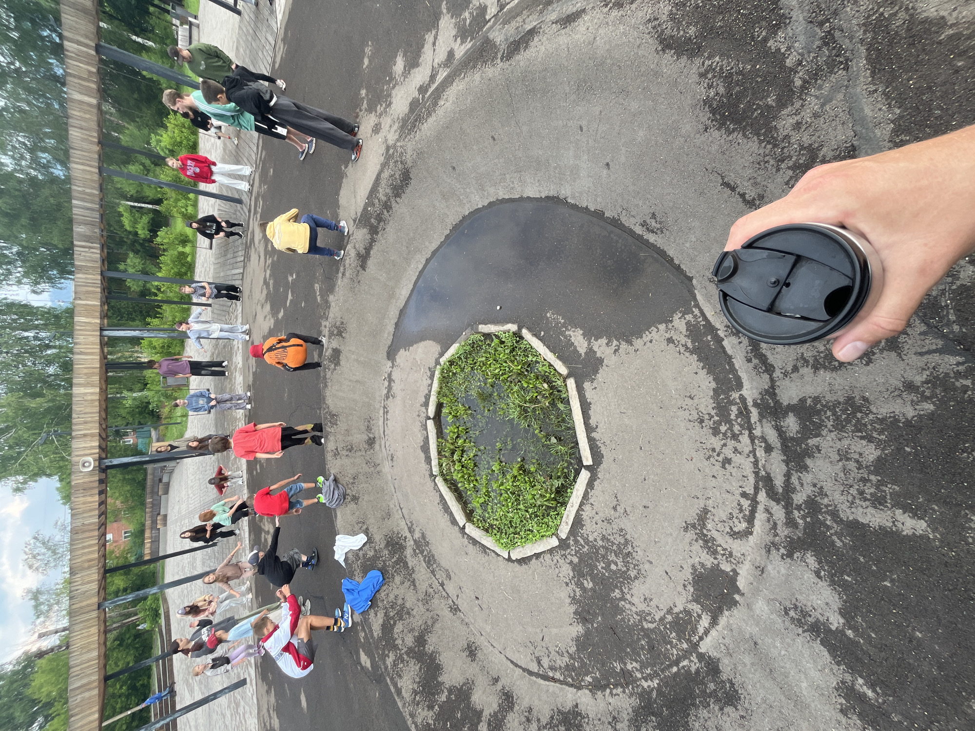 In Divnogorsk, Russia, a group of approximately 20 young people, a mix of genders and ages, are gathered outdoors in a park-like setting. They are engaged in what appears to be a coordinated physical activity, possibly a dance or exercise class, as they are arranged in a semi-circle around a central, circular, water-filled planter box with a concrete edge. Many are in motion, with some crouched, others reaching, and some standing. The environment features a large, wooden, curved structure overhead, supported by black metal poles, suggesting a covered seating or performance area. The ground is paved with asphalt and concrete, with patches of wetness and some scattered debris, including blue and white clothing items and a grey bag. Trees and foliage are visible in the background, indicating a natural or park setting. The perspective is from a slightly elevated viewpoint, looking down at the group, with a human hand holding a black and silver insulated tumbler in the foreground, partially obscuring the scene.
