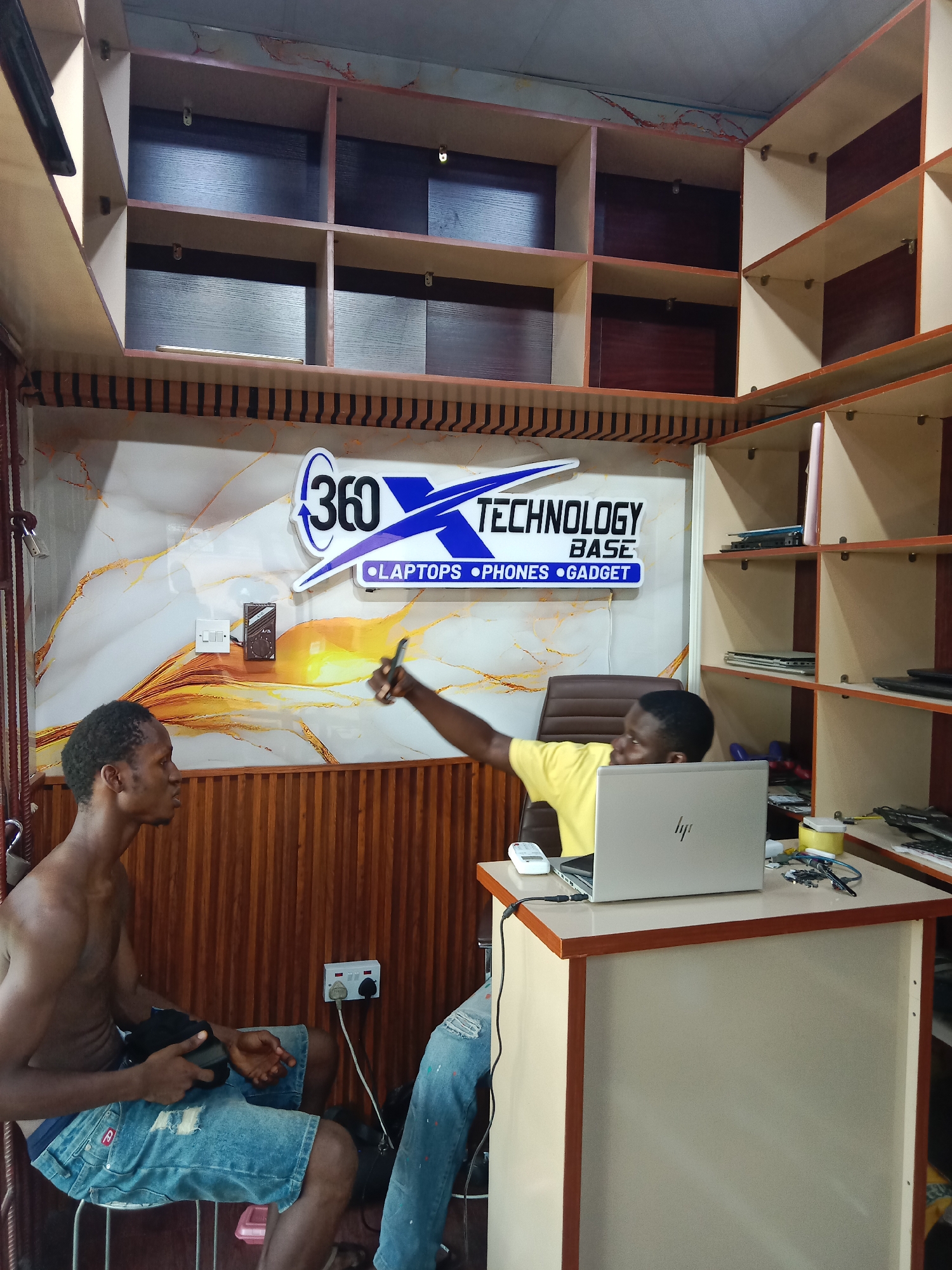 The image shows the interior of a small shop, presumably a technology store in Onitsha, Nigeria. Two men are present. One man is sitting shirtless on a stool to the left, wearing distressed denim shorts and holding a dark-colored object. The second man, wearing a yellow t-shirt, is sitting behind a small desk on which a laptop is open. The second man is extending his arm, holding a smartphone as if taking a photo of the first man. A sign behind the desk reads "360 Technology Base - Laptops - Phones - Gadget." The shop has shelves lining the walls filled with what appear to be laptops and other electronic devices. There is a wooden paneling around the walls. An electrical outlet and a small electronic device is visible on the wall next to the shirtless man.