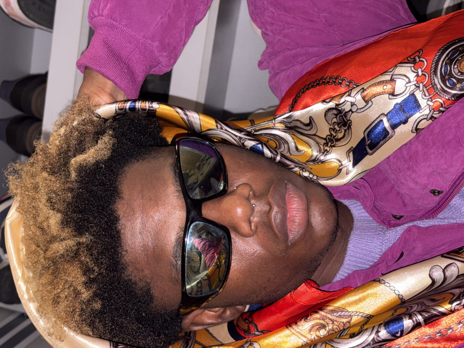 A person lies on their side, head resting on a chair back adorned with a patterned scarf featuring red, gold, and blue. Sunglasses reflect the surrounding environment. The person wears a lilac shirt and a purple jacket. Hair is dark with blonde highlights. A small piercing is visible in the nose. A rack of shoes is partially visible in the background. The entire scene is rotated counter-clockwise, presenting an unconventional perspective.