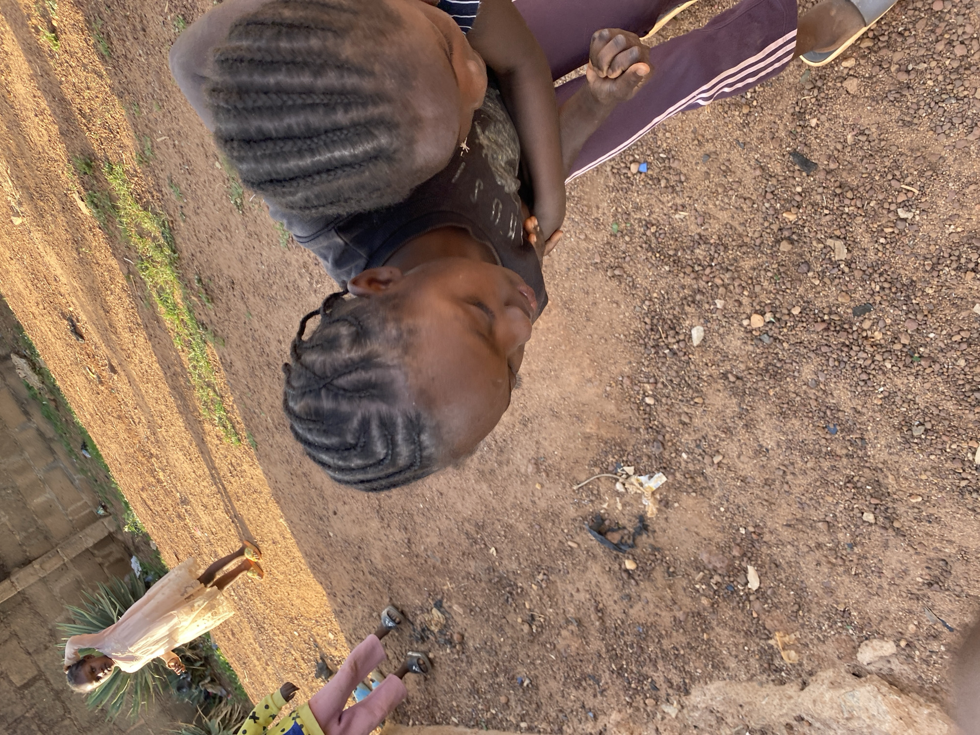 Two children are at the center of the image. One child is seated, wearing maroon pants with white stripes on the side, a dark-colored top, and tan sandals. Another child is sitting on their lap, and they are embracing. Both children have braided hair. The ground is dirt and pebbles. An individual in a light-colored dress and yellow sandals is in the background. Another person is partly visible in the bottom left corner, wearing yellow. The image may be taken in Bukuru, Nigeria.
