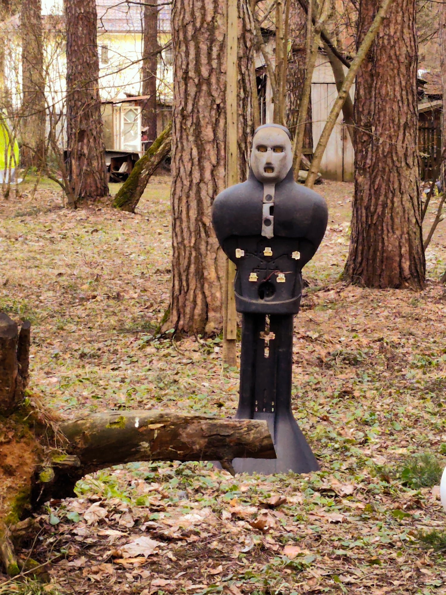 The image depicts a martial arts training dummy in a wooded area, possibly in Kratovo, Russia. The dummy is dark-colored, humanoid in shape with an open mouth and eye holes. It appears to be constructed of metal with several attachments and markings including a cross. The ground is covered in leaves and some green foliage. Trees are located around the dummy and a partially visible building can be seen through the trees. A log and tree stump are in the foreground to the left.