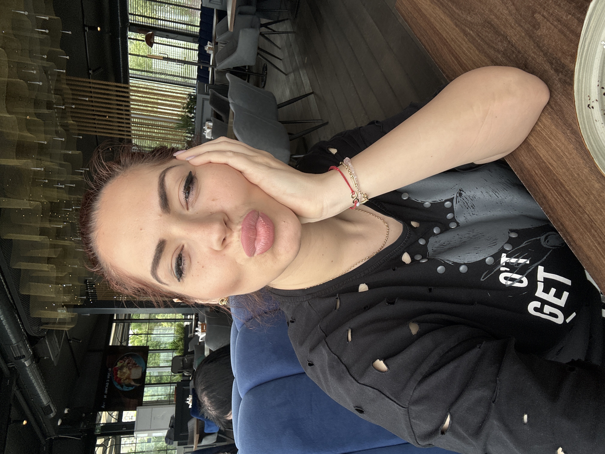 A young woman is posing in a restaurant. She is sitting in a plush blue chair with a wooden table to her right. She is wearing a black t-shirt with rips in it, and bracelets on her left wrist. She is facing the camera with her hand on her face and her lips puckered. Outside the windows of the restaurant are green trees and a view of what appears to be a city street. The woman has dark eyebrows and fair skin. The location is potentially Vidin, Bulgaria.