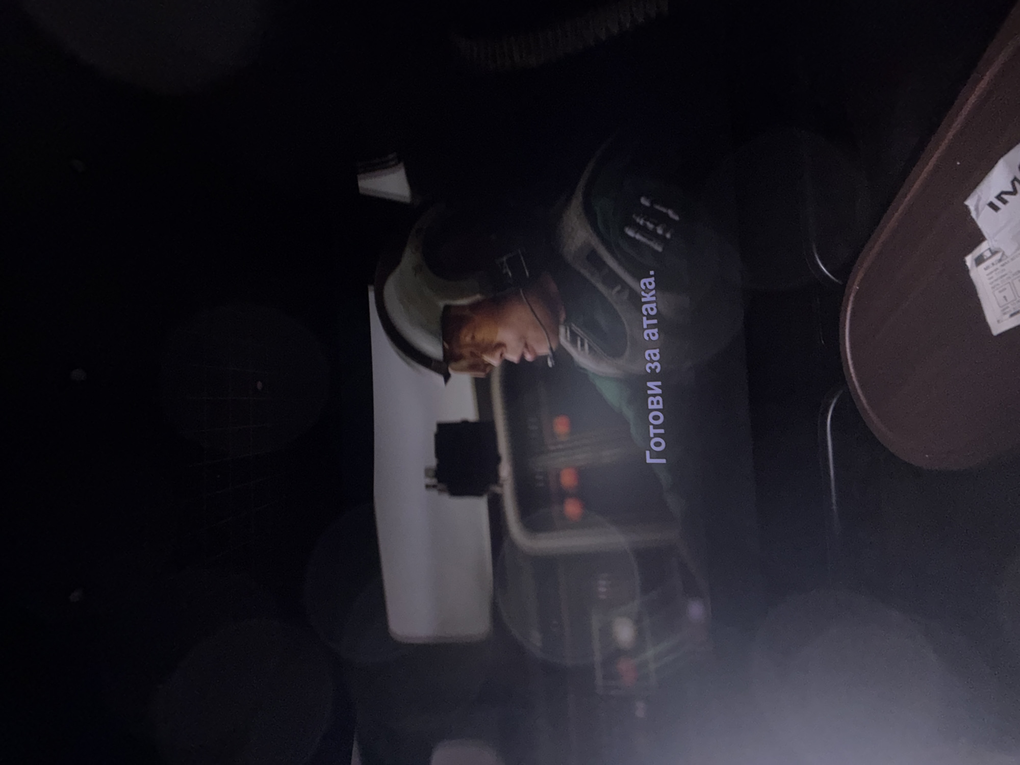 The image shows a man in a helmet, likely in a cockpit or control panel. Text "Готови за атака." is visible on the screen. There is a table on the right side. The background is dark. The location is not discernible from the image.