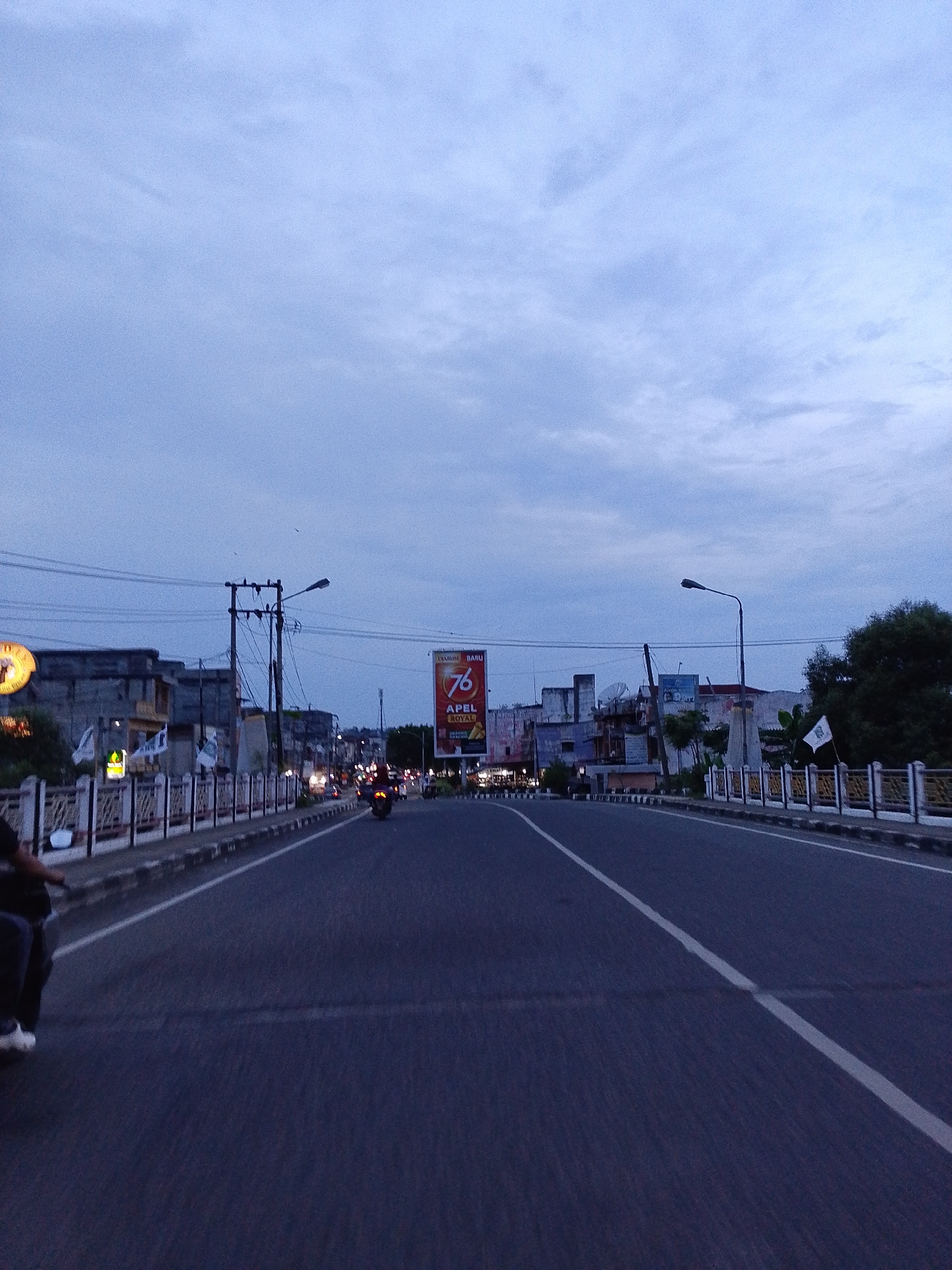 This image captures a street scene in Lhokseumawe, Indonesia, likely at dusk or dawn due to the dim, overcast sky. The wide perspective is taken from a moving vehicle, probably a motorcycle, with a person's leg and shoe visible in the lower left foreground.
The central subject is a multi-lane asphalt road with a prominent white dividing line running down the middle, extending into the distance. Decorative white and black railings or fences line both sides of the road, suggesting a bridge or elevated section. Beyond these railings, buildings of varying sizes and styles are visible, typical of an urban environment. Utility poles with numerous overhead power lines are on the left, and a streetlamp stands on the right.
Several motorcycles are present on the road; one is seen from behind further down the left lane, and others with their lights on are visible in the distance. The scene depicts routine urban commuting or travel.
Notable details include a large billboard on the right side of the road in the middle distance. It features the text "DIARUM BARU" in the top left, a large number "76", and "APEL ROYAL" below it. Small white flags are attached to the railing on the right. The sky is largely covered by uniform blue-grey clouds, indicating a lack of direct sunlight. The overall atmosphere is neutral, capturing an everyday moment in the city.