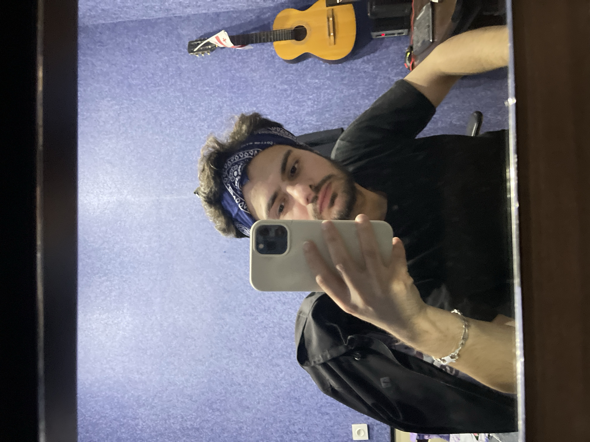 A young man with curly dark hair, a blue bandana, and a short beard looks into a mirror, taking a selfie with a white iPhone. He is wearing a black t-shirt and a black jacket draped over his left shoulder. A silver chain bracelet adorns his left wrist. Behind him, a textured blue wall serves as the backdrop. To the right, a yellow acoustic guitar with a small flag of Georgia attached to its headstock is propped against the wall. In the lower right corner, a desk is visible with a computer monitor, a keyboard, and a mouse. A power outlet is present on the wall to the left of the man. The scene appears to be set in a room within a residence, possibly a studio or bedroom, in the city of Tbilisi, Georgia.