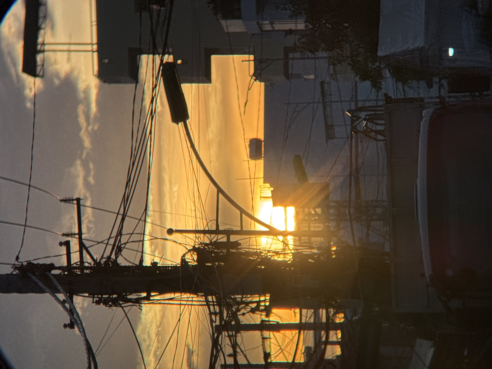 The image captures a sunset in Nha Trang, Vietnam, with the sky ablaze in hues of orange and yellow. In the foreground, a dense network of utility poles and tangled wires dominates the scene, characteristic of urban infrastructure in Vietnam. The poles are dark and weathered, supporting a chaotic web of electrical and telecommunication lines. A streetlight arm extends across the frame from a pole on the right, its dark form silhouetted against the bright sky.
Behind the utility poles, buildings rise, some appearing modern with white facades and sharp angles, while others are less distinct, blending into the background. One building has a large, flat roof structure, possibly an advertisement billboard. Another features a cylindrical tank on its roof. Sun rays pierce through the wires and the gaps between buildings, creating a dramatic lighting effect.
In the lower part of the image, the dark silhouette of a vehicle, possibly a truck or van, is visible. The scene suggests a typical urban environment in Nha Trang, where a dense population necessitates extensive utility infrastructure, set against the backdrop of a vibrant sunset. The image conveys a sense of everyday life in a bustling Vietnamese city.