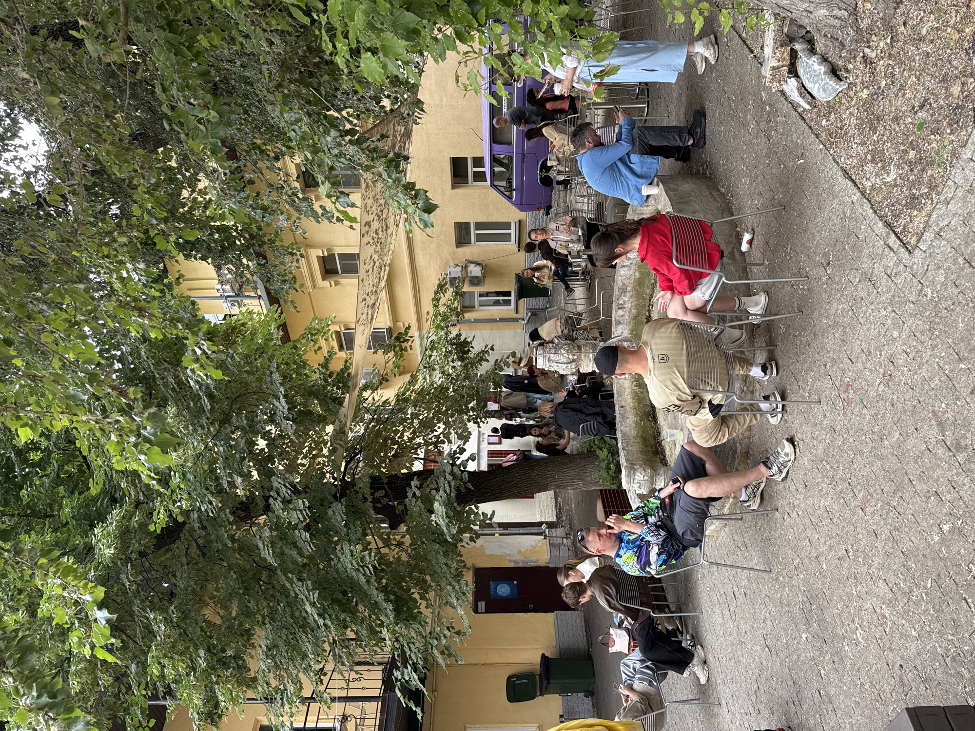 A group of people are seated at tables in an outdoor courtyard. The courtyard is paved with cobblestones and surrounded by a yellow building with windows. Trees with green leaves provide shade. A purple van is parked in the background. Some people are drinking and talking, while others are looking at their phones. There is a green trash can on the left side of the image. The scene appears to be a casual gathering at an outdoor cafe or restaurant.