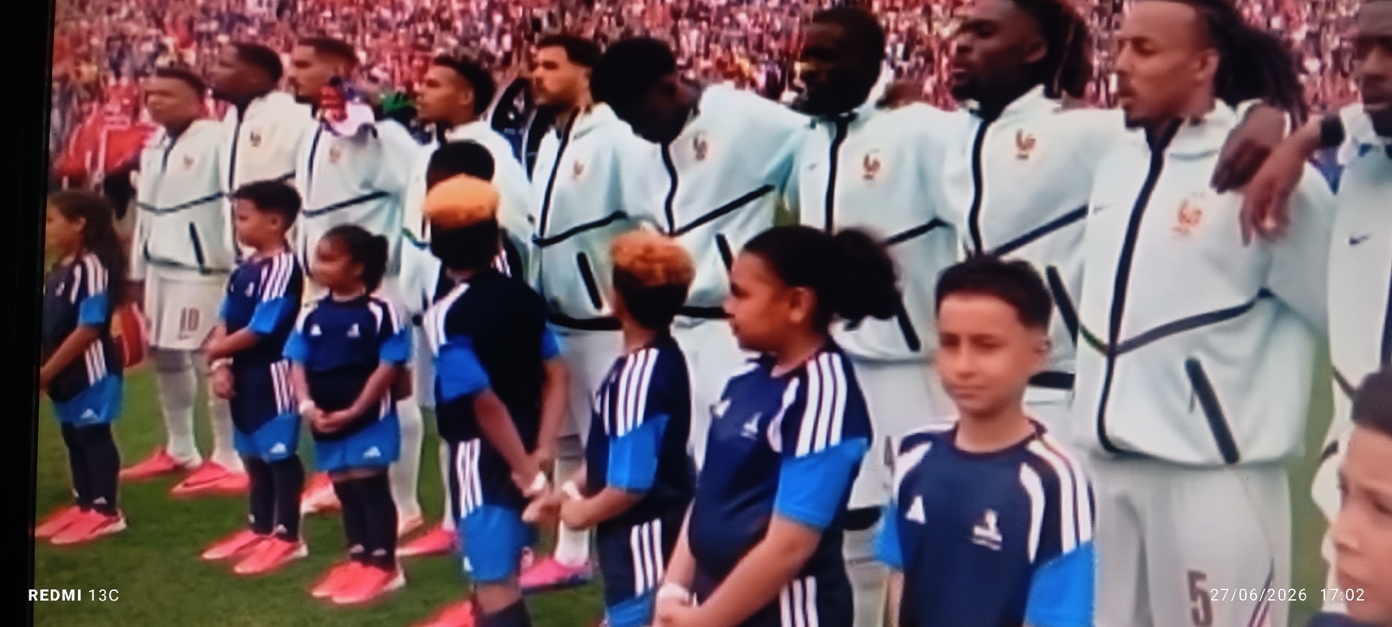A group of children and adults, likely soccer players, stand in a line on a grassy field in front of a stadium crowd. The children in the foreground are wearing matching blue and white soccer uniforms. The adults behind them are wearing white tracksuits with blue accents and appear to be part of a national team, as indicated by crests on their jackets. The crowd in the background is blurred but suggests a sporting event. The overall atmosphere appears to be one of anticipation or a pre-game ceremony. There is no information in the image to suggest the location is the city of Idimu, Nigeria.