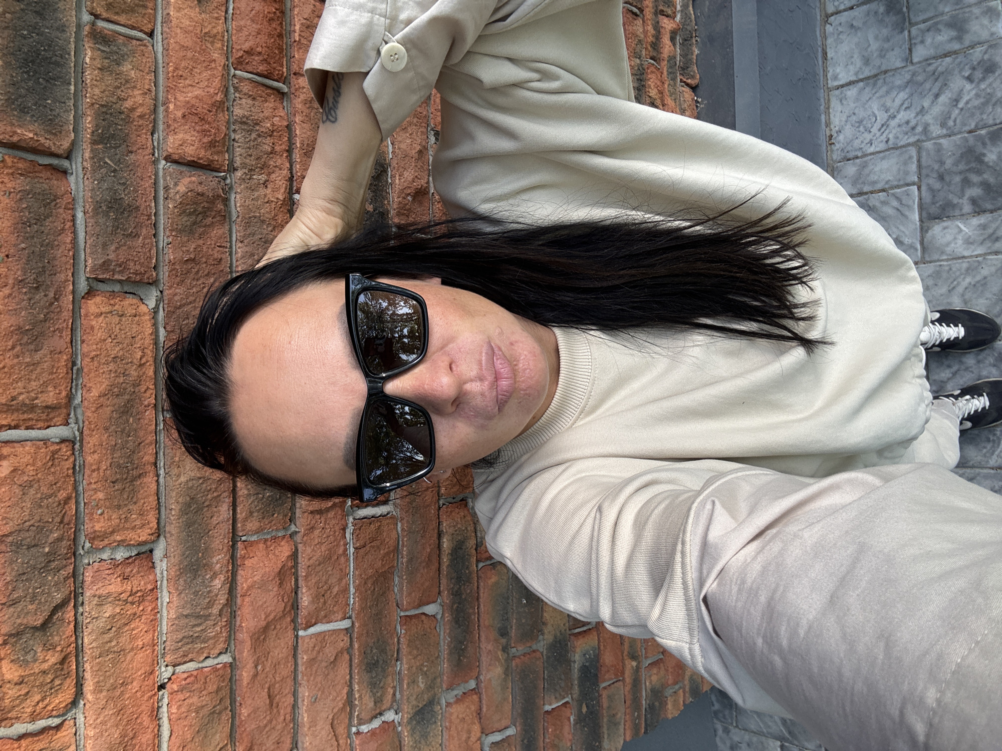 A person wearing dark sunglasses is positioned close to a brick wall. The wall is made of red and brown bricks with light-colored mortar. The person is also wearing a light-colored, long-sleeved shirt with a button on the sleeve, and light-colored trousers. The person's hair is dark and falls across the shirt. The background includes gray pavement and part of a black and white shoe. The location is Chaikovsky, Russia.