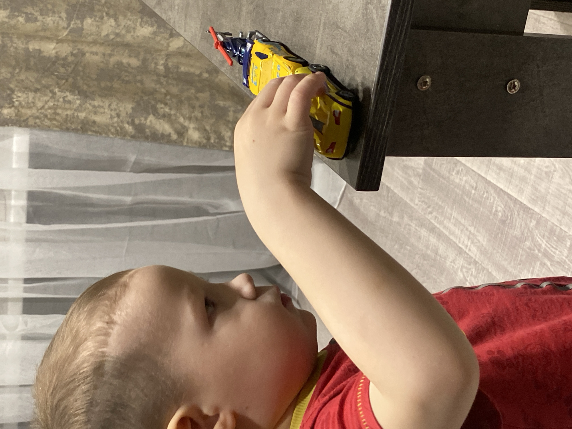 An indoor scene depicts a young child, visible in profile from the chest up, facing right. The child has short, light-colored hair and wears a red t-shirt with a yellow collar trim and yellow stitching visible on the right sleeve. The child's right hand is positioned over a yellow toy sports car, which rests on a dark, flat rectangular surface, likely a table or shelf. The sports car has "G-CO" and other text on its side. Attached to the rear of the sports car is a second yellow toy vehicle, appearing to be a tow truck or flatbed, carrying a small blue and silver toy helicopter with a red main rotor blade. The furniture piece supporting the toys features visible screw heads on its vertical panel. In the background, sheer white curtains are present to the left, suggesting a window. To the right of the curtains, a wall is covered with textured wallpaper in shades of brown, beige, and gold. The floor is a light-colored laminate or tile with a visible wood grain pattern. The specific location within the city of Sochi, Russia, cannot be determined from the provided image.