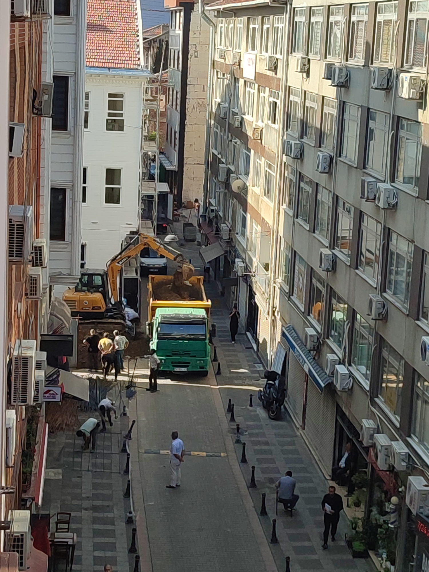 The image depicts a narrow city street in Kadıköy, Turkey, with buildings lining both sides. Construction work is underway in the middle of the street. A yellow excavator is actively digging, with its bucket depositing soil into the back of a green dump truck. Several construction workers, dressed in casual clothing, are present at the scene, some observing the work and others appearing to be involved in it. One worker, wearing a light blue shirt and white pants, stands near the dump truck, seemingly overseeing the operation. Further down the street, other individuals are visible, including people walking and sitting, suggesting it is a public thoroughfare. The buildings on either side of the street are multi-story structures with numerous windows and air conditioning units installed on their facades. The street is paved with cobblestones.