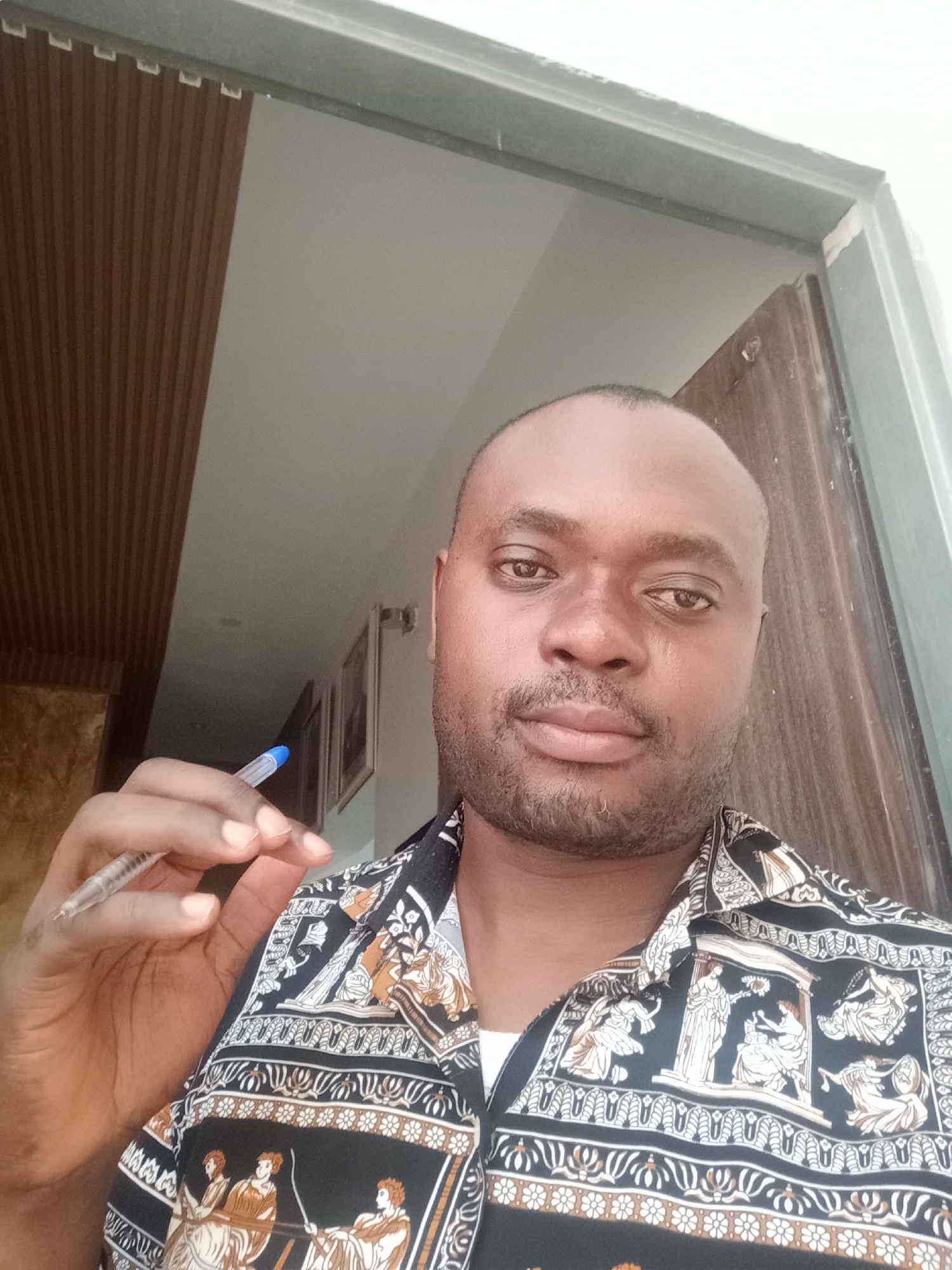 A man, appearing to be in his 30s or 40s, is captured in a medium close-up, eye-level selfie. He has a dark complexion, a shaved head with very short dark hair, and a neatly trimmed beard. He is wearing a black button-up shirt featuring intricate white, gold, and brown patterns that depict classical figures and decorative borders. In his right hand, he holds a clear blue ballpoint pen between his thumb and forefinger, positioned to the left of his chest. He looks directly at the camera with a neutral to slightly serious expression.
The background indicates an indoor setting, likely within a residence or office in Gwarinpa, Nigeria. To the man's left, a section of a wall is covered with vertical brown wooden slats. To his right, part of a light green-grey door frame and a dark brown wooden door or panel are visible. The upper left background shows a plain light-colored wall, possibly white or beige, with some blurry framed pictures hanging further back. The lighting is even and consistent with an interior during daytime. No discernible text is visible on the shirt patterns or the pen itself.