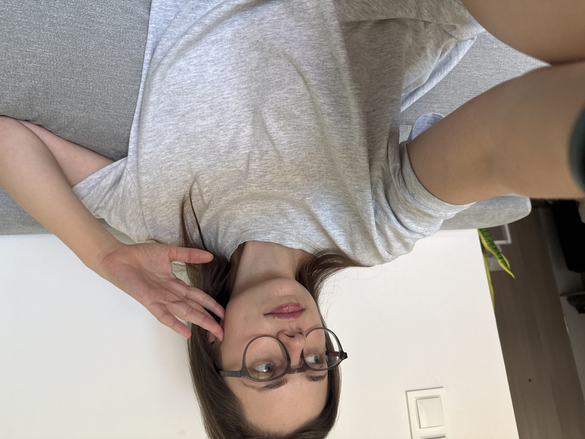 A young woman with long brown hair and wearing glasses and a grey t-shirt is sitting on a grey couch. She is looking to the left, with her right hand resting on her cheek. The background is a plain white wall with a light switch. A potted plant with green leaves is visible in the background to the left. The scene is indoors, in what appears to be a living room. There is no indication of the city of Szklarska Poręba, Poland.