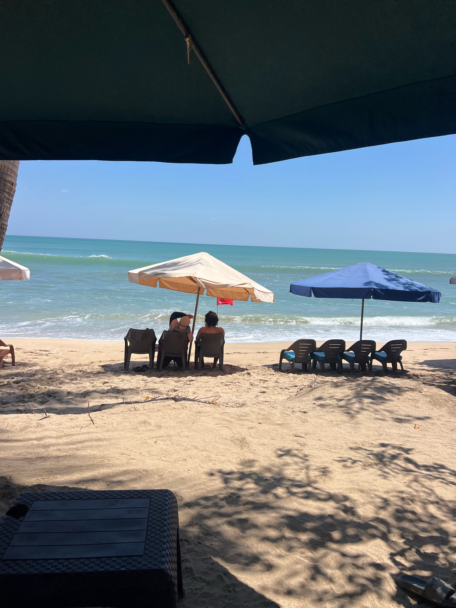 A beach scene in Kuta, Indonesia, shows a sandy shore with the ocean in the background. Two beach umbrellas, one beige and one dark blue, are set up on the sand. Under the beige umbrella, three people are seated in chairs, facing the ocean. Under the blue umbrella, four empty chairs are arranged in a row. A dark-colored, textured coffee table sits in the foreground, partially in shadow. The water is a turquoise color with gentle waves. The sky is clear and blue. Shadows from palm trees and foliage are cast across the sand.