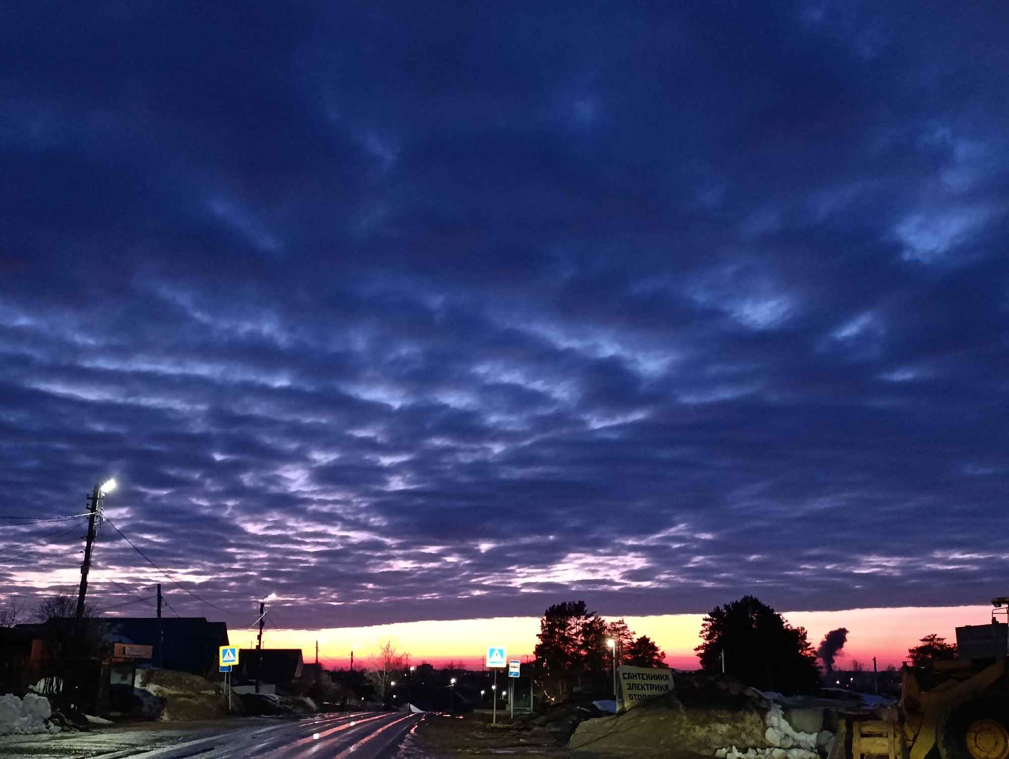 The image depicts an outdoor street scene in the city of Chaikovsky, Russia, at dawn or dusk. The sky, occupying the upper two-thirds, features extensive, horizontally stratified altocumulus or similar cloud formations ranging from dark blue-purple at the zenith to lighter purple and a prominent band of orange-pink at the horizon. The lower third of the image shows a wet or icy asphalt road curving slightly to the right, reflecting illuminated streetlights. Patches of snow are visible on the ground along both sides of the road.
On the left, a tall utility pole with multiple power lines supports an illuminated street light. Behind it, dark silhouettes of low-rise buildings are discernible. A blue-and-yellow pedestrian crossing sign stands near the road's edge.
On the right, another blue-and-yellow pedestrian crossing sign is visible further down the road, accompanied by additional illuminated streetlights. A cluster of dark coniferous trees stands in the midground. A prominent white sign, facing the viewer, displays Cyrillic text which translates to "PLUMBING ELECTRICITY HEATING." To the far right, a portion of a yellow heavy machinery, possibly a front-end loader, is partially visible, positioned near a mound of snow or earth. In the distance, against the colorful horizon, a distinct plume of dark smoke rises, suggesting an active chimney or industrial source. No individuals are present in the scene.