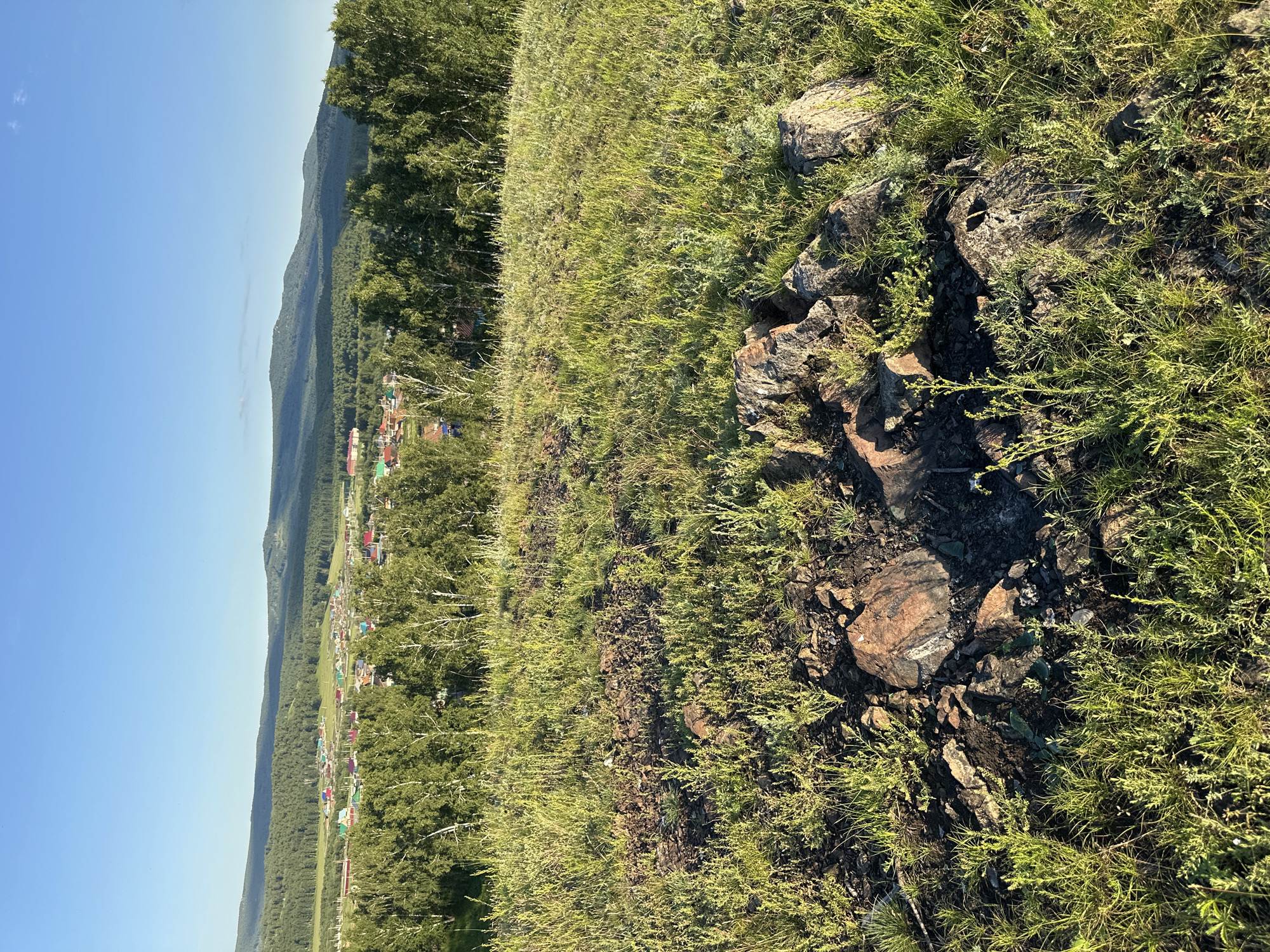 This media file captures a natural landscape scene with a human settlement visible in the distance. The foreground is dominated by a grassy hill with scattered rocks and sparse vegetation. The grass is green and appears to be under bright sunlight.
In the mid-ground, a cluster of buildings, possibly a village or small town, is nestled amidst a dense forest. The buildings have various colored roofs, suggesting human habitation. The forest stretches out behind the settlement, leading to rolling hills or mountains in the background. The sky is a clear, bright blue, indicating a sunny day.
The image is oriented vertically, with the sky occupying the left side and the landscape filling the right. The lighting suggests it is daytime, likely late morning or early afternoon due to the angle of the sun casting shadows. The overall mood is peaceful and serene, highlighting the coexistence of nature and human settlement. No people or specific objects are clearly identifiable in the foreground or mid-ground. No text is visible in the image.