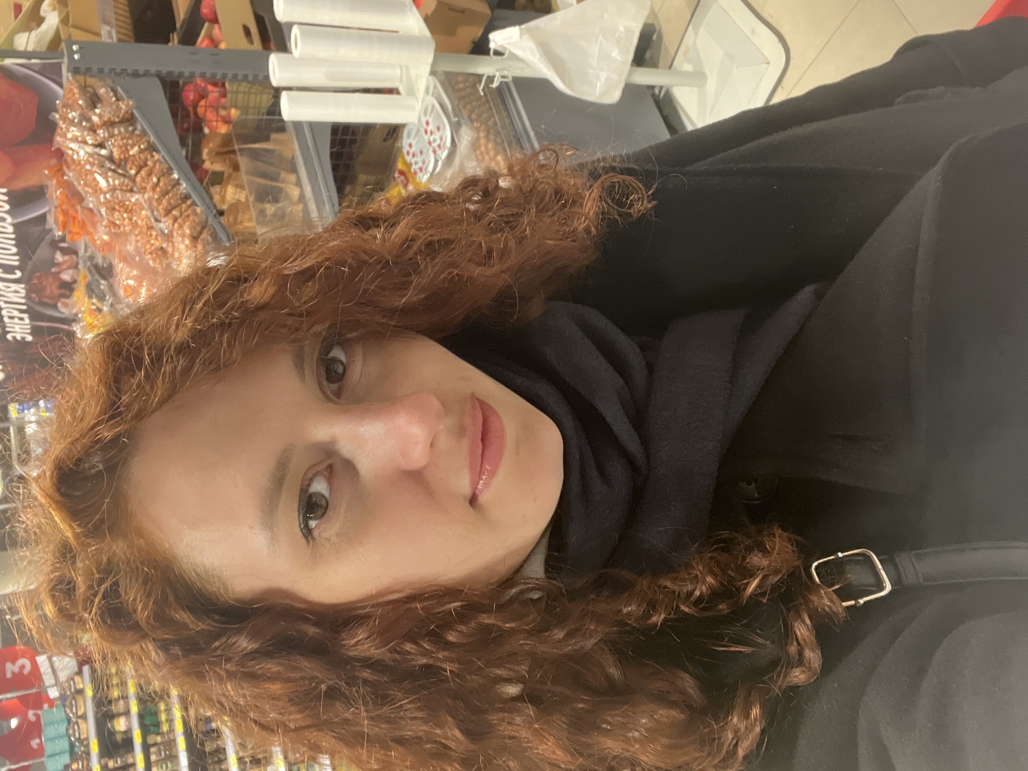 The image displays a close-up portrait of a woman, presented on its side, in what appears to be a grocery store or supermarket in Saint Petersburg, Russia. The woman has voluminous, reddish-brown curly hair that frames her face. She looks directly towards the viewer with a neutral to slightly smiling expression, her lips slightly parted. She is wearing a dark scarf, likely black or navy, and a black coat or jacket. A metallic buckle, possibly from a bag strap or coat detail, is visible near her right shoulder.
In the background, typical store shelving is visible, stocked with various products. Packaged goods, including what look like nuts or dried fruits in clear bags, are on display. Red fruits, possibly apples, are also discernible. White cylindrical objects, likely part of the shelving system, are present. On an orange or red background in the store, partial Cyrillic text can be seen, which reads "ЭНЕРГИЯ С ПОЛНОЙ..." (ENERGIYA S POLNOY...), suggesting an advertisement or product description related to energy. The lighting is artificial, indicative of an indoor setting.
