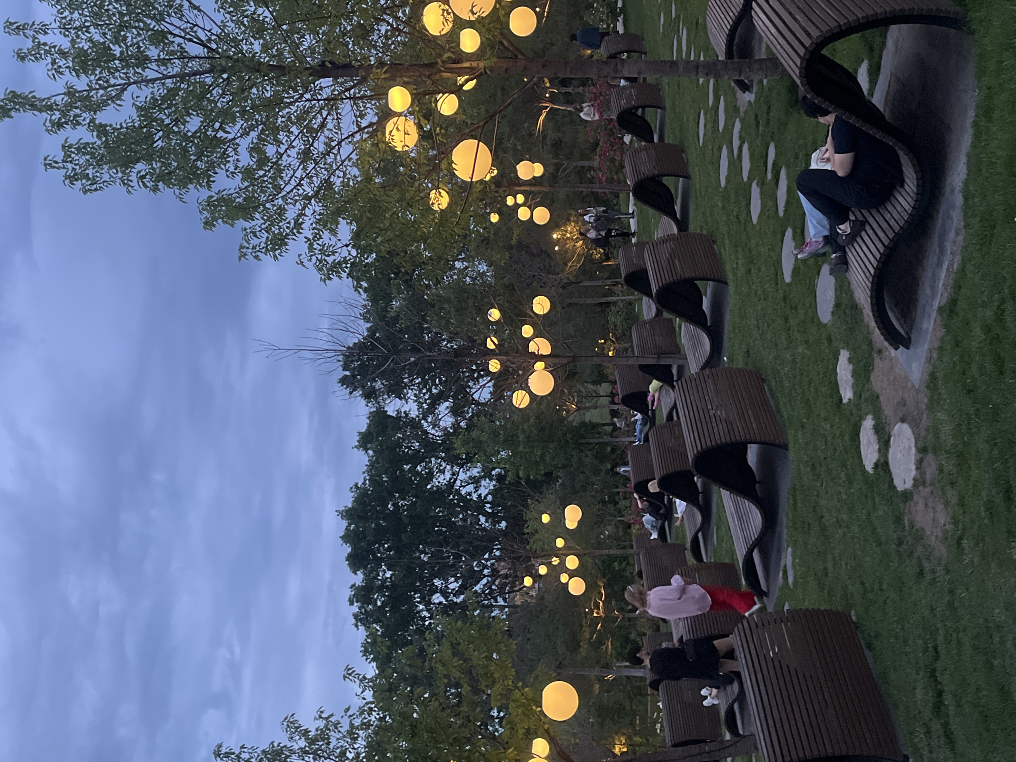 The image captures an evening scene in a public park or garden, likely in Krasnodar, Russia, under a twilight sky. Numerous spherical, yellow-orange lanterns are suspended from the branches of lush green trees, casting a warm glow across the area.
A series of distinctive, curved wooden benches with slatted designs are arranged along a path on the right side of the frame. Several individuals are present, with some seated on these benches, and others walking or standing further in the background. One person on a bench in the foreground is wearing a black top and light blue jeans, while another in the mid-ground is dressed in a pink top and red pants. The ground features a mix of green grass and irregularly shaped light-colored paving stones that form sections of the pathway.
The soft blue-gray sky indicates dusk or early evening, with the artificial lighting from the lanterns creating a serene and inviting atmosphere. People appear to be engaged in leisure activities, enjoying the tranquil setting. No text is visible in the image.
