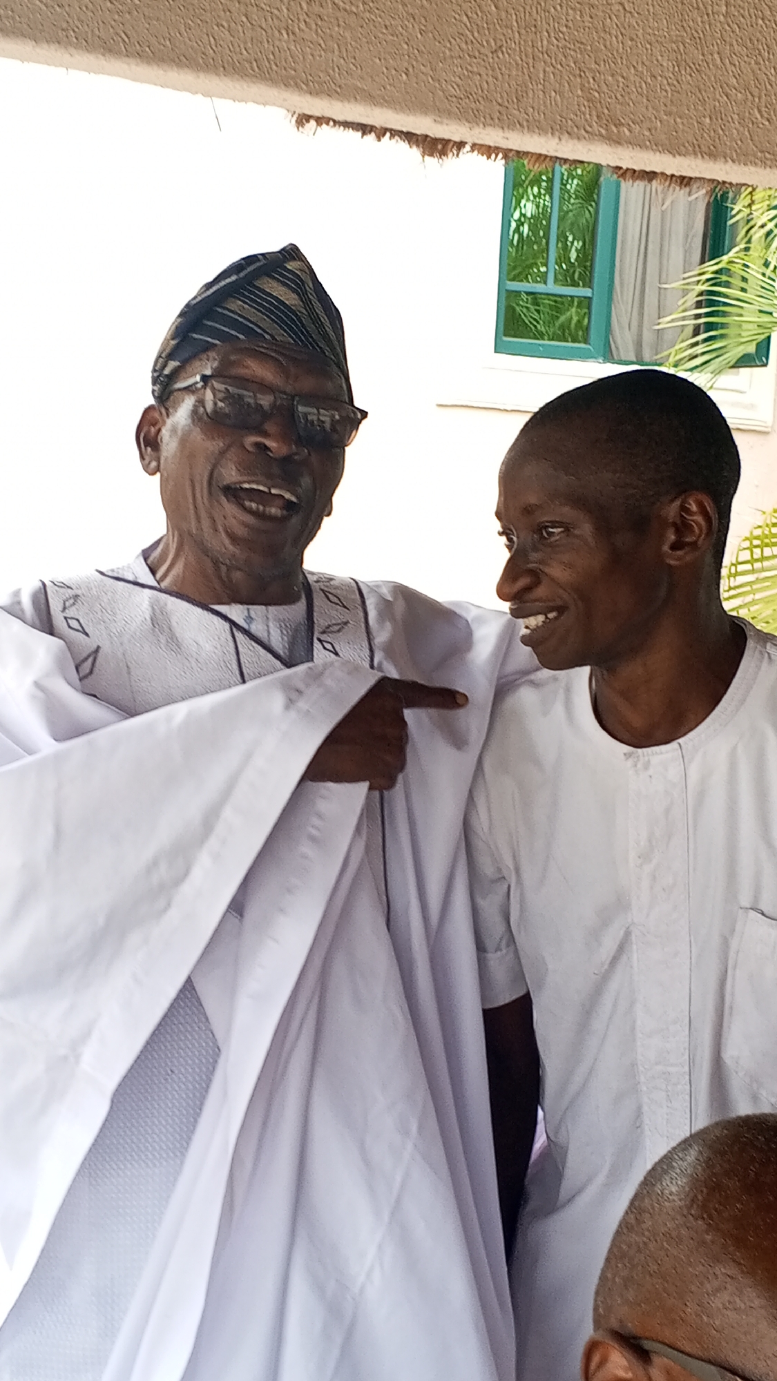 Two men are standing together, smiling and interacting. The man on the left is older, wearing a traditional patterned hat and glasses, and a white robe-like garment with dark trim. He is pointing with his right hand towards the other man. The man on the right is younger, wearing a plain white shirt, and is looking at the older man with a smile.
The background is a building with a window; part of a green plant is visible outside the window. The location is potentially Lokoja, Nigeria, based on the assumption from the prompt.