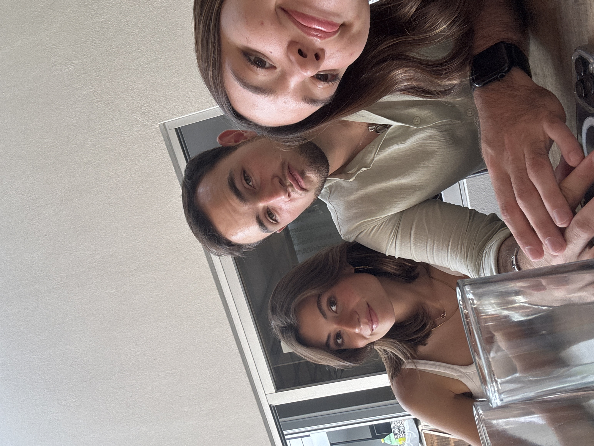 A selfie-style photograph depicts three individuals sitting at a table, presumably indoors. A woman with long brown hair is at the top of the frame, a man with short dark hair and a beard is in the middle, and another woman with shorter brown hair is at the bottom of the frame. The man wears a light green collared shirt, and the women are wearing light colored tops. A watch is visible on the arm of the man, who appears to be holding a phone. In the foreground, stacked glass objects are visible. Beyond the individuals, a window or glass door is visible, suggesting an indoor setting. The location is undetermined and cannot be identified from the photograph. It cannot be determined from the image that this location is in the city of San Bernardino Tlaxcalancingo, Mexico.