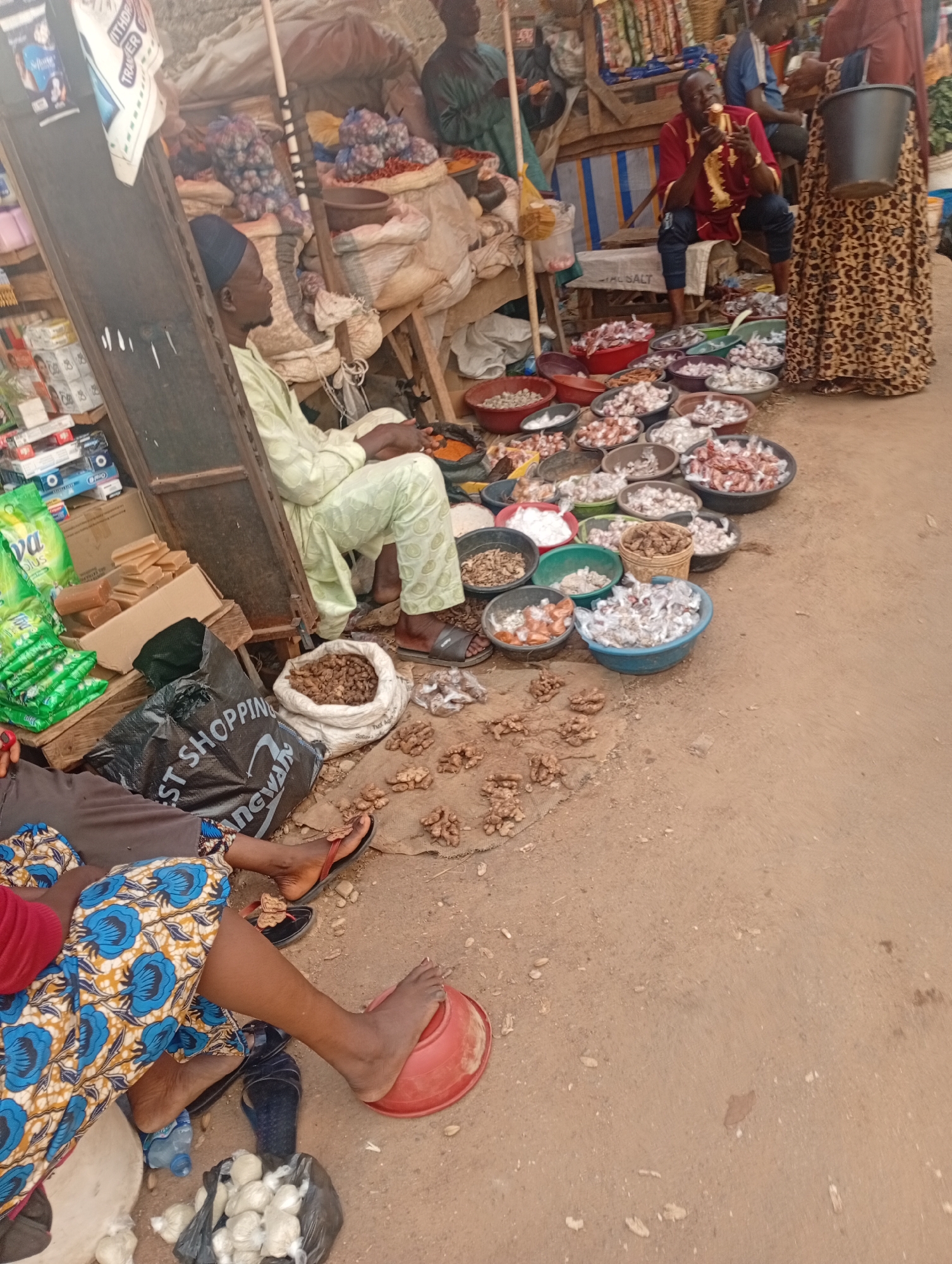 A busy outdoor market scene is depicted, likely in Rafin Par, Nigeria. Several individuals are present, engaged in market activities. In the foreground, a person wearing a vibrant blue and yellow patterned fabric sits, with one foot resting on a red bowl. Nearby, a black shopping bag with white lettering, partially visible as "ST SHOPPING," is on the ground. Another person in a light green traditional garment sits with their hands clasped, facing away from the camera, positioned next to a wooden stall filled with goods.
Further back, a man in a red embroidered shirt sits on a bench, holding a piece of food to his mouth. He is surrounded by rows of bowls containing various items, likely spices or dried goods. A woman in a patterned skirt stands beside him, holding a dark bucket. The market stall displays an assortment of products, including bags of what appear to be onions or garlic, sacks of dry goods, and numerous bowls filled with small items, possibly herbs, spices, or dried seafood.
The ground is covered in dirt and sand, indicative of an outdoor market. The lighting suggests it is daytime, possibly morning or afternoon. The overall atmosphere appears active and commercial, with people involved in selling and purchasing goods. The presence of traditional attire and the general market setup provide strong cultural and geographical cues.