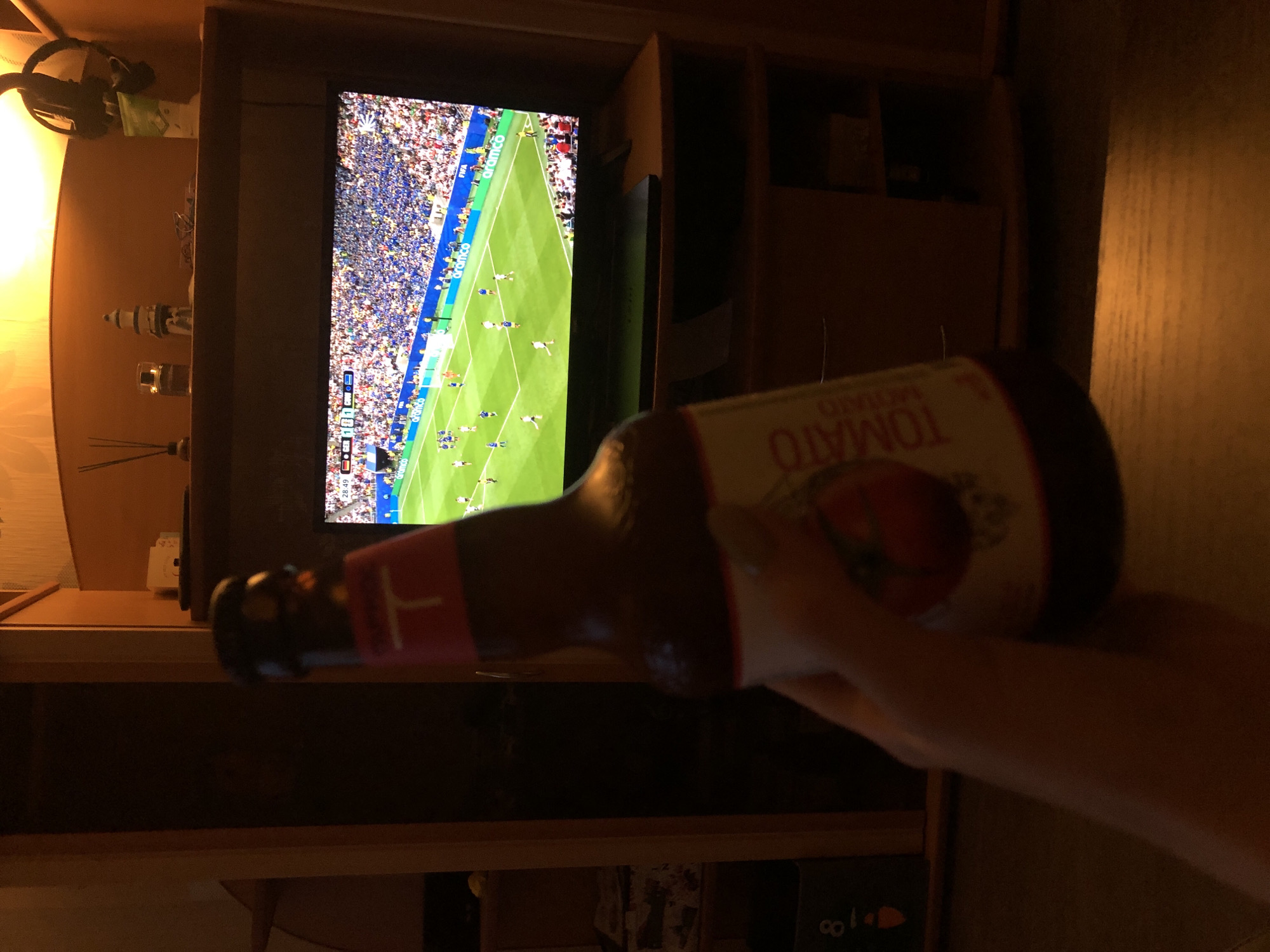 In a dimly lit room, a person's hand holds a bottle of "TOMATO" beer in the foreground, obscuring part of a television screen. The television displays a soccer match with players in blue and white uniforms on a green field. The stadium appears full of spectators, mostly in blue. The room is furnished with wooden cabinets and shelves, and a lamp casts a warm glow from the top right corner. A diffuser with sticks and a small lighthouse figurine are visible on a shelf above the television. The environment suggests a home entertainment setting, likely in Chaikovsky, Russia.