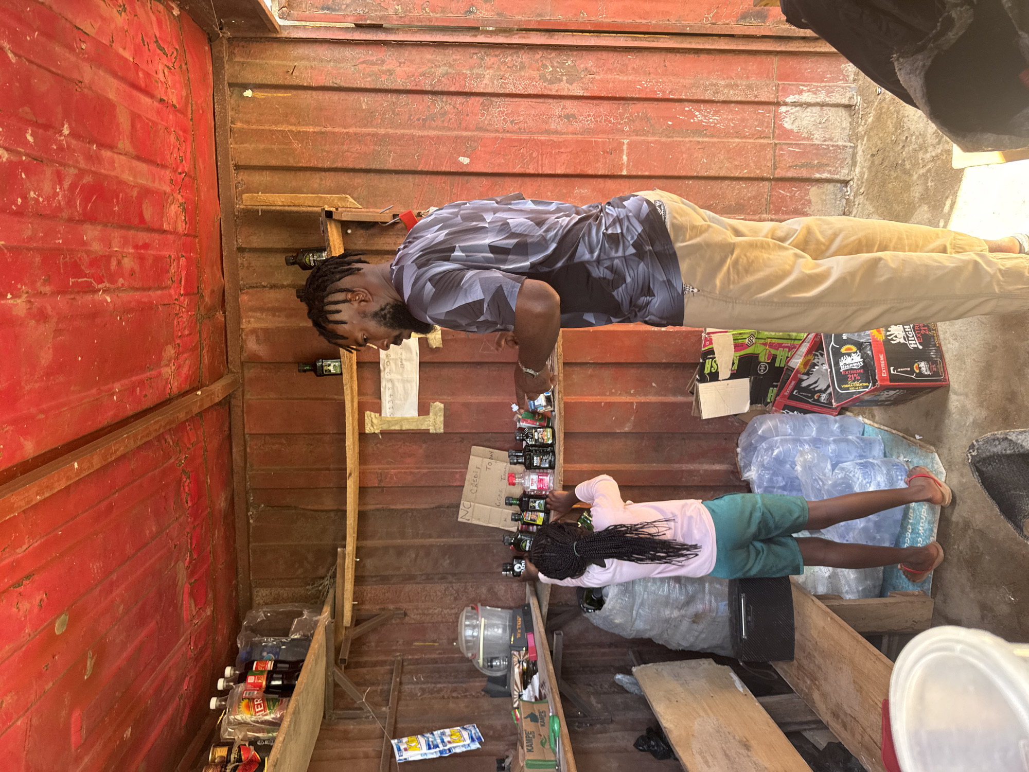 In a roadside stall in Jos, Nigeria, a young girl with braided hair reaches for a bottle on a shelf, while a man with dreadlocks stands nearby, observing. The stall, constructed from corrugated metal, has a red ceiling and walls. Shelves are stocked with various bottled items, some in plastic packaging, and a sign reads "NO CREDIT TO DAY." Water gallons are stacked on the floor, alongside boxes and bags. The scene captures a moment of commerce within a simple, utilitarian setting.