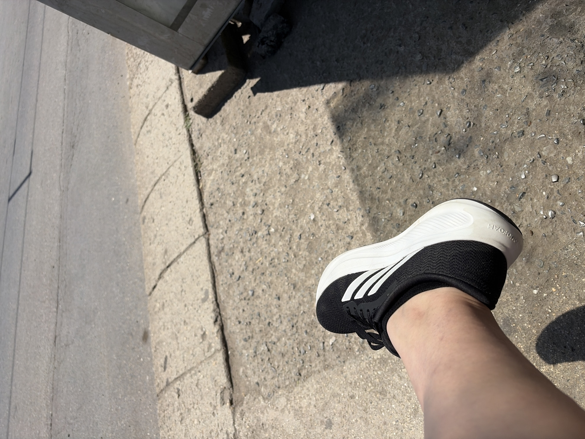 A person's lower leg and foot, wearing a black and white Adidas sneaker, rests on a gritty, concrete surface. The shoe has three white stripes on the side and a white sole. The concrete appears to be a sidewalk or pathway, with distinct grout lines visible and scattered small stones and debris. In the background, a paved road surface is visible, with subtle markings suggesting lanes. The scene is illuminated by bright sunlight, casting a distinct shadow from the person's foot and shoe onto the ground. To the right, a portion of a wooden structure, possibly a sign or barrier, is visible, propped up by concrete blocks. The location is identified as Stara Zagora, Bulgaria.