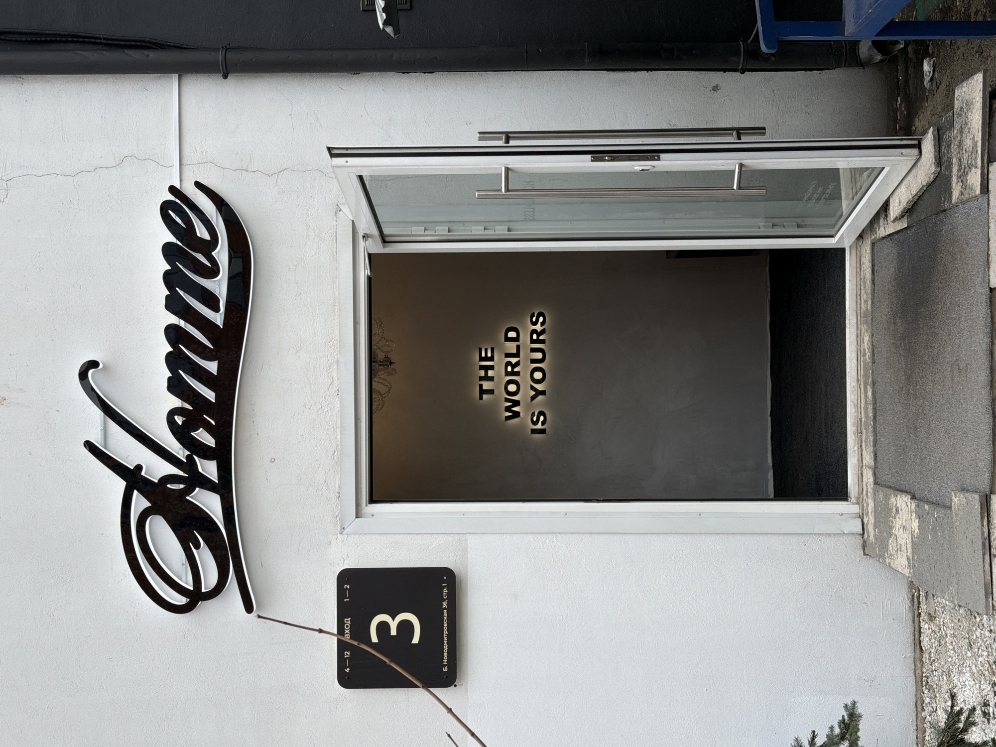 A photograph captures the entrance of a business located at Б. Новодмитровская 36, стр. 1 in Khimki, Russia. The building features a textured, off-white stucco facade with several vertical hairline cracks visible, particularly to the right of the door.
Prominently displayed above the entrance is a three-dimensional black cursive sign with a white outline spelling "Homme." To the left of the door, a rectangular black plaque displays white text: "4-12 ВХОД 1-2" at the top, a large numeral "3" centrally, and "Б. Новодмитровская 36, стр. 1" along the bottom. A slender, bare tree branch extends diagonally across the upper left portion of this plaque.
The entrance comprises a modern white-framed glass door, which is open inward at approximately a 45-degree angle. The door is fitted with two vertical, silver-toned bar handles. The visible interior beyond the threshold appears as a dark gray, carpeted room. On the back wall of this room, illuminated text glows with the phrase "THE WORLD IS YOURS." A faint reflection of a decorative light fixture or chandelier is visible on the interior ceiling.
At ground level, directly in front of the open door, lies a dark gray rectangular floor mat. Beyond this, a series of uneven, light-colored concrete or stone slabs form a path, transitioning to an area of white and light gray gravel on the left. A small evergreen shrub is partially visible in the lower-left corner of the frame. To the right of the entrance, a black vertical downspout or pipe is affixed to the building, and a section of a blue, bench-like structure is visible on the far right edge of the image. No individuals are present in the scene. The ambient lighting suggests an overcast or muted daytime condition.
