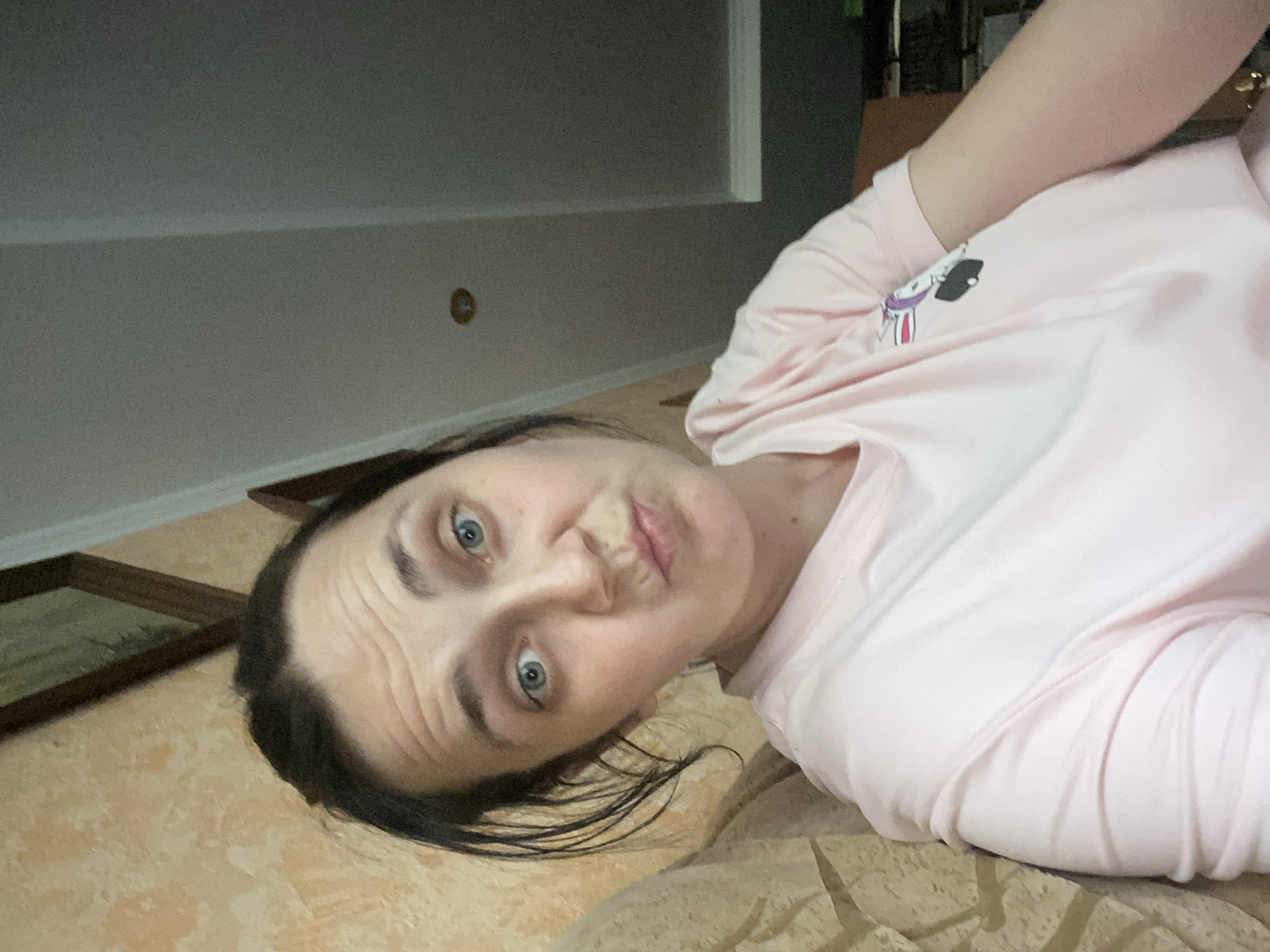 A woman with dark hair pulled back, blue eyes, and fair skin makes a puckered-lip expression at the camera. She is wearing a light pink t-shirt with a graphic of a rabbit riding a skateboard on the left side of her chest. She is leaning against a textured, light brown wall. The background shows a slanted ceiling with recessed lighting.