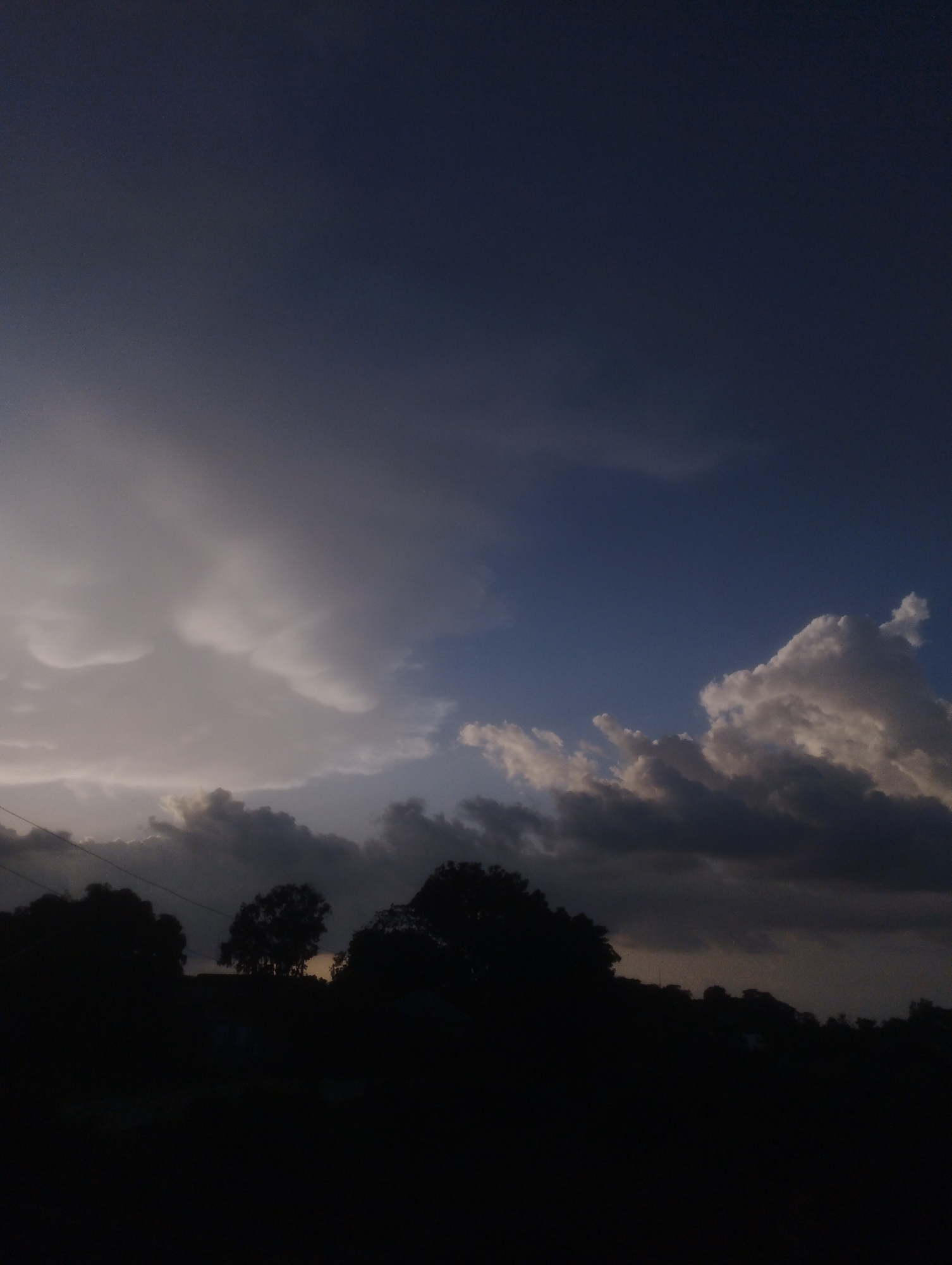The image depicts a twilight sky over Jos, Nigeria, characterized by dramatic cloud formations and silhouettes of trees and vegetation against the fading light. The sky transitions from a deep, dark blue at the top to lighter shades of blue and grey towards the horizon. Several distinct cloud types are visible: wispy, lighter clouds are seen towards the left, while darker, more ominous storm clouds gather on the right and in the mid-ground. The lower portion of the image is dominated by the dark silhouettes of trees and a slight hint of distant landforms, obscuring any ground-level details. The overall impression is one of an approaching or receding storm, creating a somber and atmospheric scene. There are no people or active events visible in the frame.