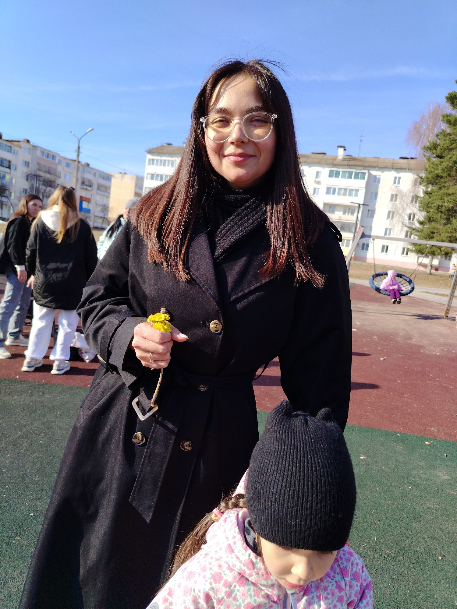 The image shows a woman in a black coat standing in a playground, possibly in Noviy Urengoy, Russia given the urban backdrop of Soviet-era apartment buildings. She is holding yellow flowers in her right hand. She is wearing glasses and has long, dark hair. A child wearing a black hat and pink floral patterned jacket is partially visible in the foreground, suggesting an interaction. In the background, other people are visible, some appear to be walking, and a child is swinging on a playground swing. The weather appears to be sunny. The playground surface is a combination of red and green.