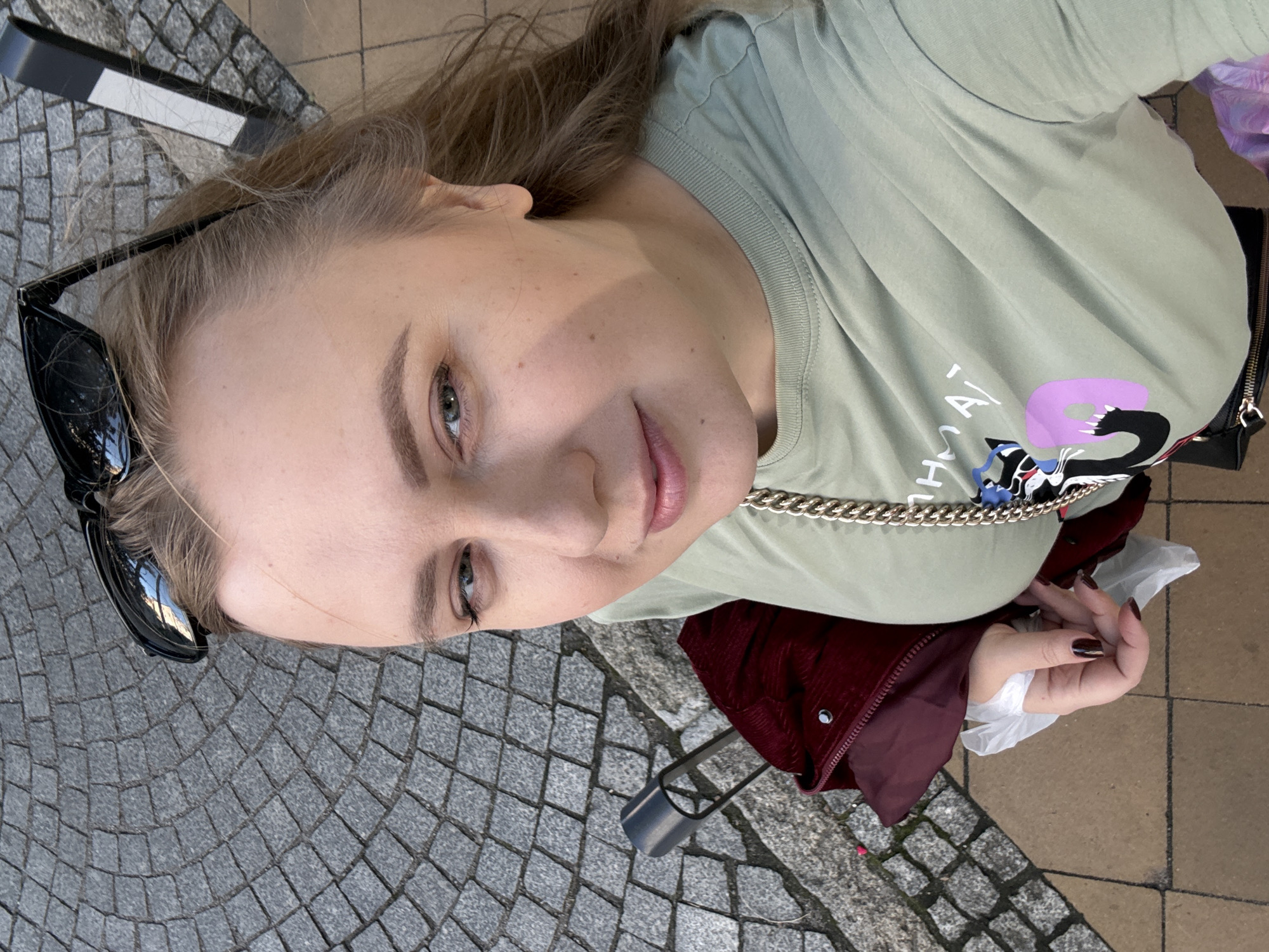 A woman is taking a selfie outdoors, with cobblestones and yellow pavement visible in the background. She is wearing a light green t-shirt with a graphic print of a black cat and pink shapes. A dark red jacket is draped over her shoulder, and a thick gold chain hangs around her neck. Her sunglasses are pushed up on her head. The scene appears to be in Zelenogradsk, Russia.