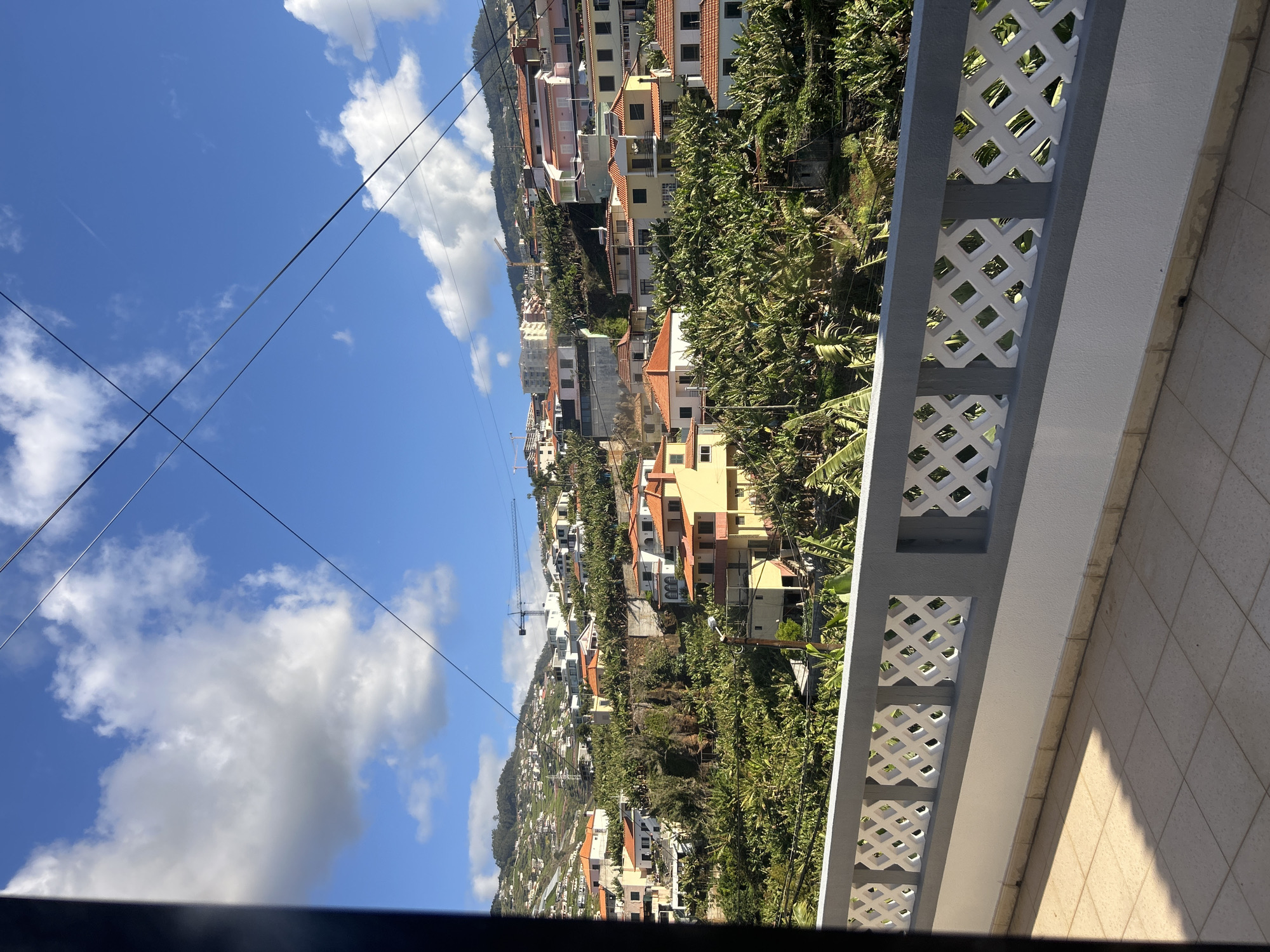 The image captures a view from a balcony in Câmara de Lobos, Portugal. The balcony has a gray and white lattice-patterned railing. Beyond the railing, a hillside covered in houses and vegetation slopes upward. The houses are various colors, with orange tiled roofs. Numerous banana trees are visible. A construction crane is seen amidst the buildings. Power lines stretch across the blue sky, where scattered white clouds are present. The balcony surface is tiled. A dark shadow is visible at the very bottom of the frame.