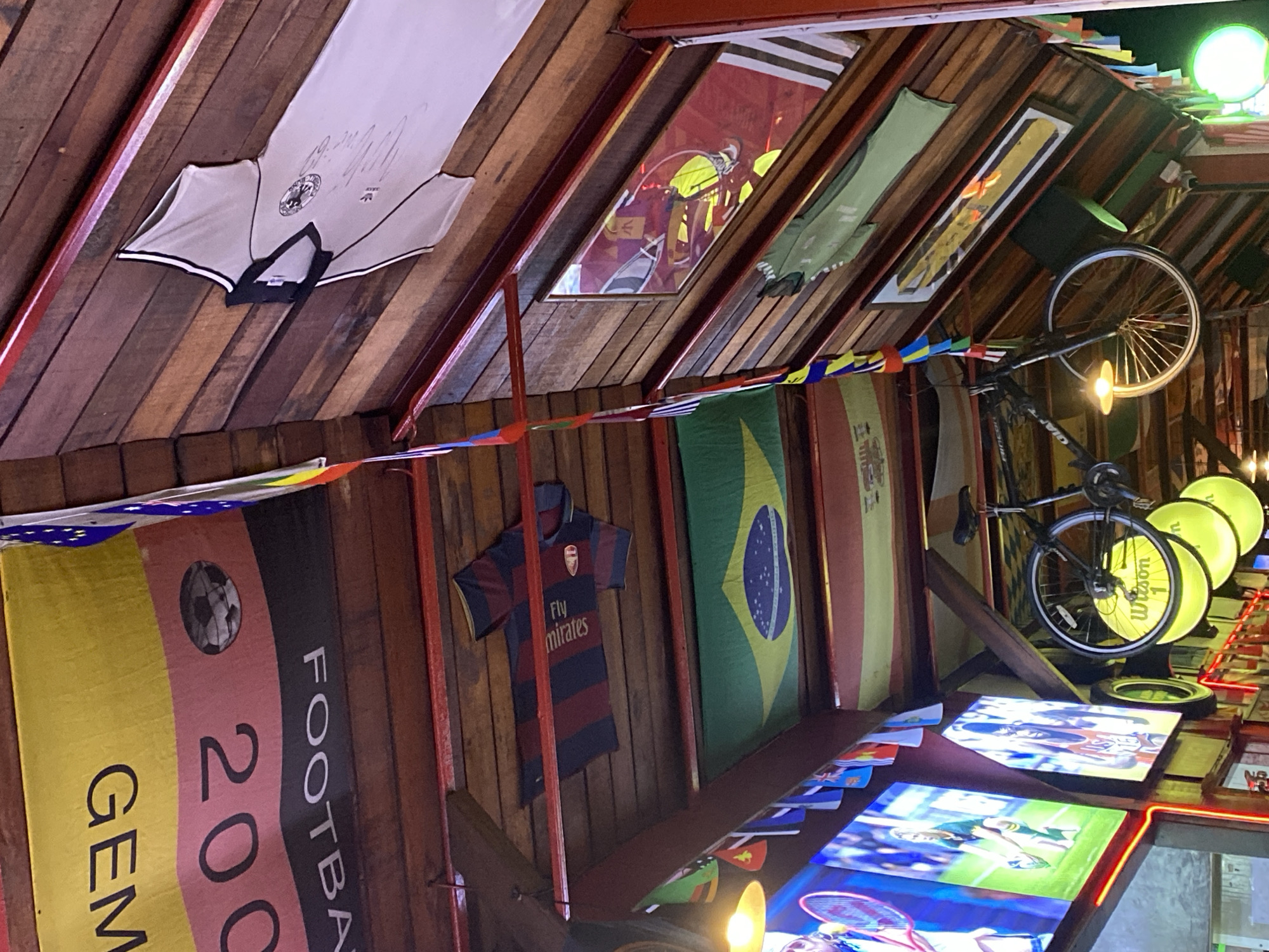 The interior of a bar in Moo 1, Thailand, is decorated with sports memorabilia. Hanging from the wooden ceiling are framed sports jerseys, flags representing various countries (Germany, Brazil), and a bicycle. Below these, televisions are mounted, displaying sports footage. A banner commemorating the 2006 FIFA World Cup held in Germany is also visible. The scene suggests a sports-themed bar catering to an international clientele.