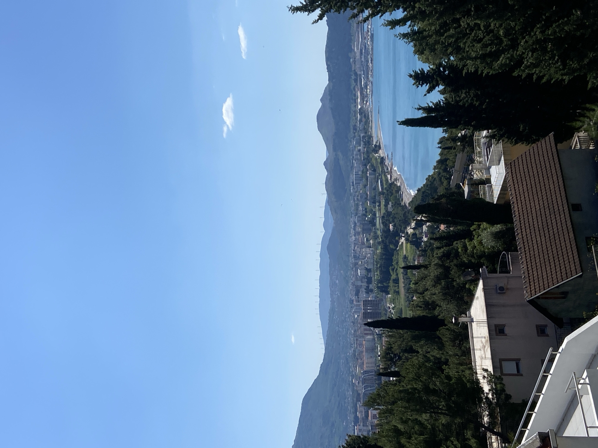 The image shows a view from Šušanj, Montenegro, overlooking the Adriatic Sea and the city. The sky is mostly clear blue with a few small clouds. In the middle ground, there are mountains partially covered with greenery and buildings. The sea is visible along the right side. In the foreground, there are trees and portions of buildings with visible rooftops and balconies.