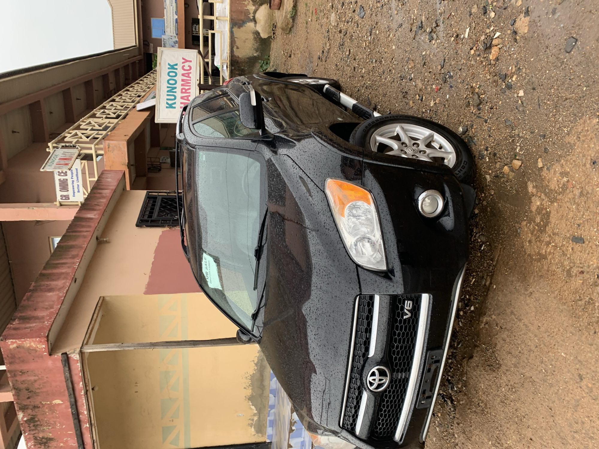 An image, rotated on its side, captures a black Toyota RAV4 V6 SUV parked on a wet, gravelly, and muddy surface next to a multi-story building in Rafin Par, Nigeria. No people are visible in the scene.
The car, covered in raindrops, suggests recent wet weather under an overcast sky. A white "FOR SALE" sign with a partially obscured phone number (08**********) is taped to the inside of the windshield. The Toyota emblem and "V6" badge are clearly visible on the front grille. The SUV's wheels feature a silver, five-spoke design, and the tires are branded BFGoodrich.
The building to the left of the car has light peach-colored walls and appears to be commercial, with several signs attached to its facade. One prominent sign reads "KUNOOK PHARMACY" in green text. Another, partially visible sign below it, says "GR OMINO CE..." with the tagline "Empowering People Better Livelihood." The building features balconies or ledges with a reddish-brown trim, showing some signs of wear and rust. A metal-grated window is visible on the lower level. The ground around the vehicle is uneven, composed of dirt, small rocks, and puddles.