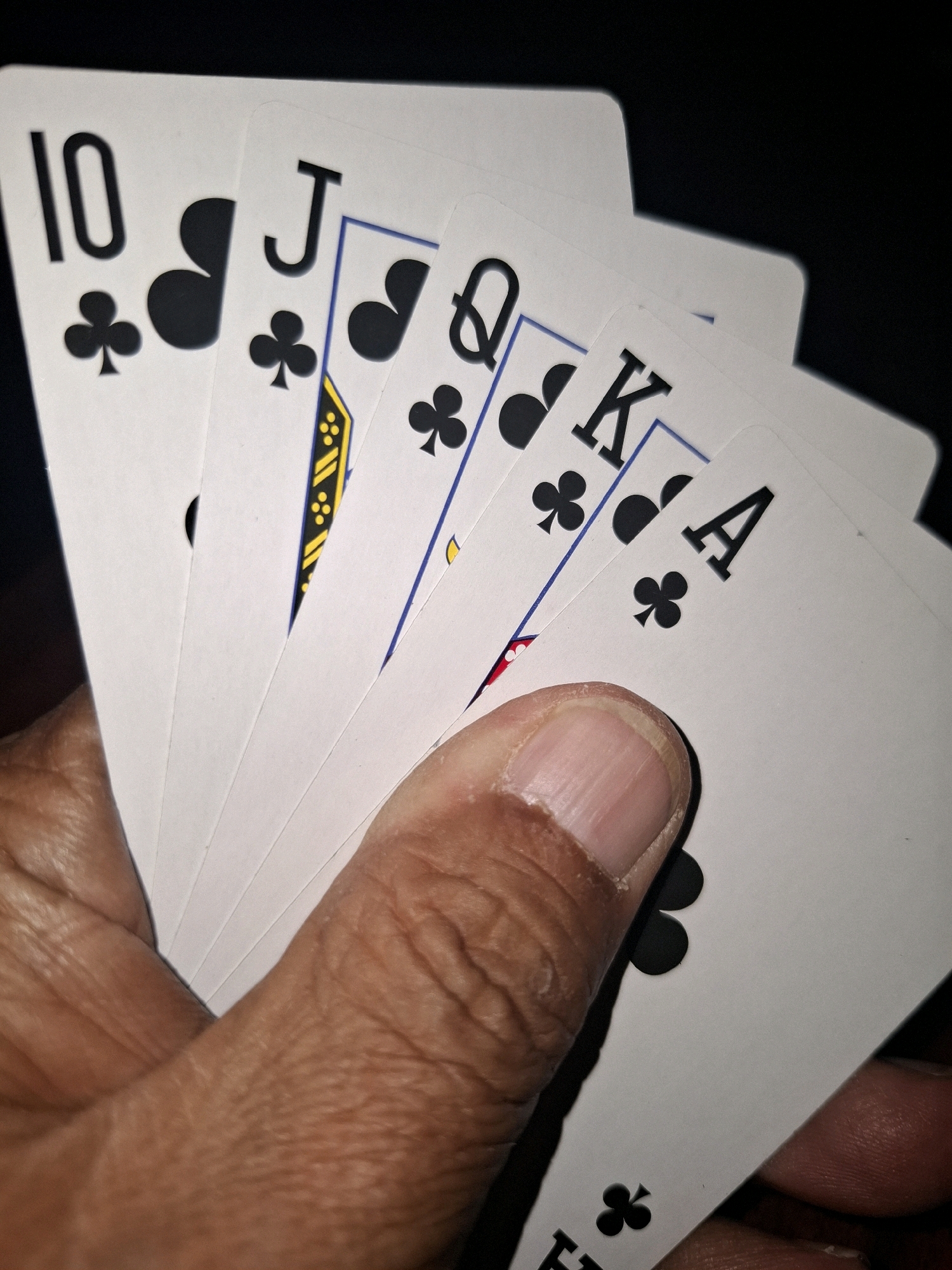A hand holds several playing cards, fanned out against a dark background. The cards visible are the 10 of clubs, Jack of clubs, Queen of clubs, King of clubs, and Ace of clubs. The lighting is dramatic, casting shadows and highlighting the textured surface of the hand. The scene is indoors, with no discernible elements to suggest a specific city or country, including Almaty, Kazakhstan.
