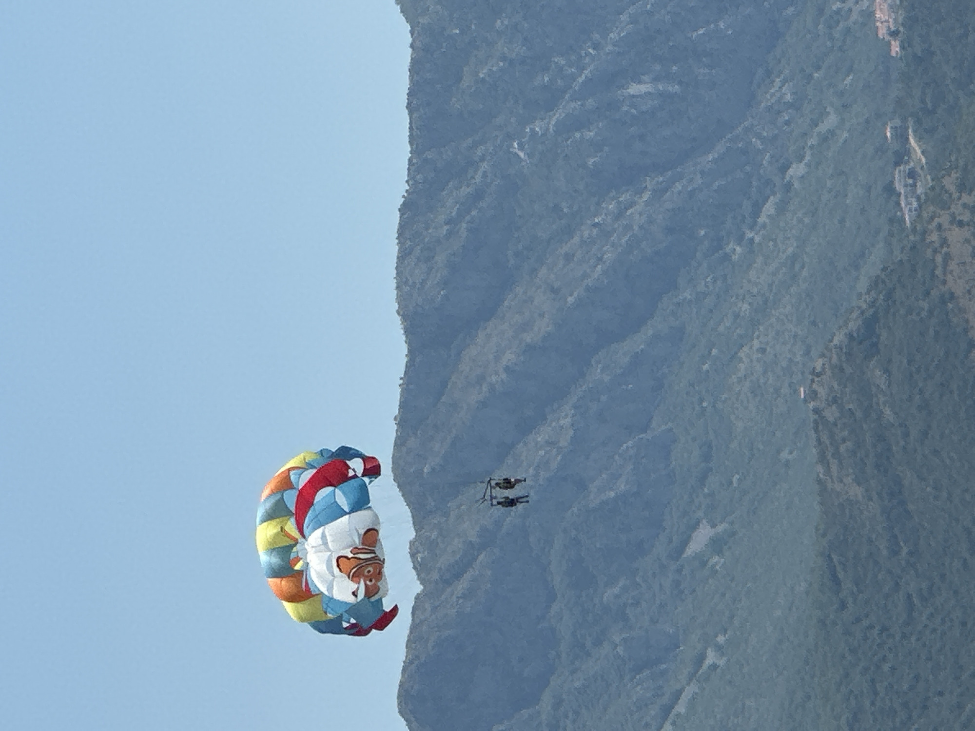 The image shows a parasailer with a colorful parachute, featuring a cartoon character, descending against a backdrop of a lush green mountainside under a clear blue sky. Two people are visible in the parasail harness, suspended beneath the canopy. The parasail is positioned in the foreground, to the left of the frame, while the mountains occupy the right and center portions. The sky is a vibrant, cloudless blue, suggesting it is daytime with good weather. The mountains are densely covered with vegetation, with some rocky outcrops visible. The scene appears to be a recreational activity, likely taking place at a coastal or mountainous tourist destination, consistent with the provided location context of Boreti, Montenegro. The overall impression is one of leisure and adventure in a scenic natural environment. No text is visible in the image.