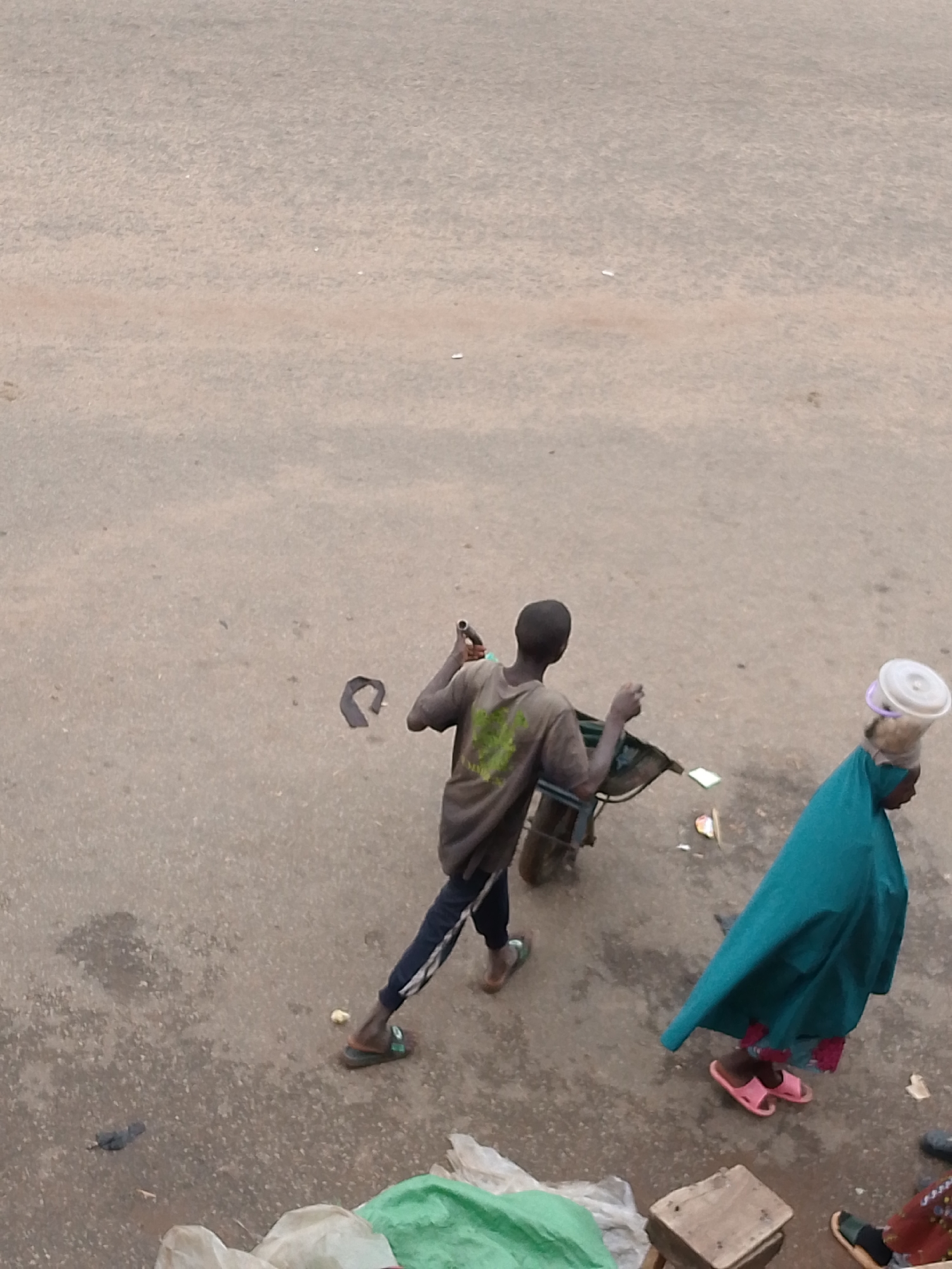In Bukuru, Nigeria, an outdoor scene depicts two individuals in a dusty, worn-down urban environment. The foreground is dominated by a rough, mottled asphalt surface, with scattered debris including plastic bags, a piece of crumpled paper, and a small yellow object. To the left, a person, likely a male, is seen from the back, wearing a grey t-shirt with a green graphic and dark pants with white stripes. He is pushing a handcart. To his right and slightly ahead, a person, likely a female, is seen in profile, wearing a teal-colored headscarf and a patterned fabric visible at the bottom of her attire. She has a white plastic basin or container balanced on her head and is wearing pink sandals. The overall impression is one of a typical street scene in a bustling, developing urban area, possibly a market.