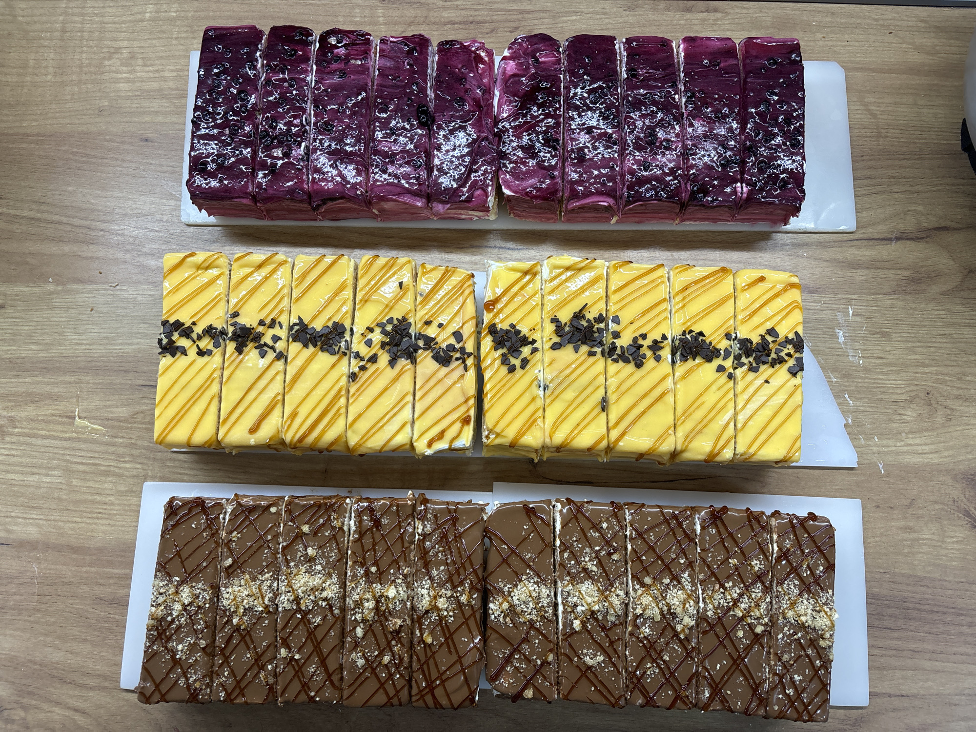 The image displays three rows of beautifully arranged rectangular desserts on a light-colored wooden surface, likely a display counter in a bakery or patisserie in Malki Iskar, Bulgaria. Each row features two long cakes, each precisely cut into multiple individual slices, presented on white rectangular trays.
The top row showcases cakes with a rich, glossy purple glaze, possibly berry-flavored, speckled with small dark fruit pieces and swirled with lighter, almost white marbling. The middle row features cakes with a vibrant yellow glaze, elegantly drizzled with caramel-colored lines, and sprinkled with dark chocolate shavings or chips along the center. The bottom row presents cakes with a smooth, deep chocolate glaze, adorned with a crisscross pattern of darker chocolate drizzles and generously topped with crushed nuts or cookie crumbs.
No people are visible, and the scene captures the desserts in a static display. The consistent cutting and symmetrical arrangement suggest careful preparation for sale or serving. The lighting is even, highlighting the colors and textures of the glazes and toppings. There is no visible text in the image. The overall impression is one of tempting and well-crafted confectionery.