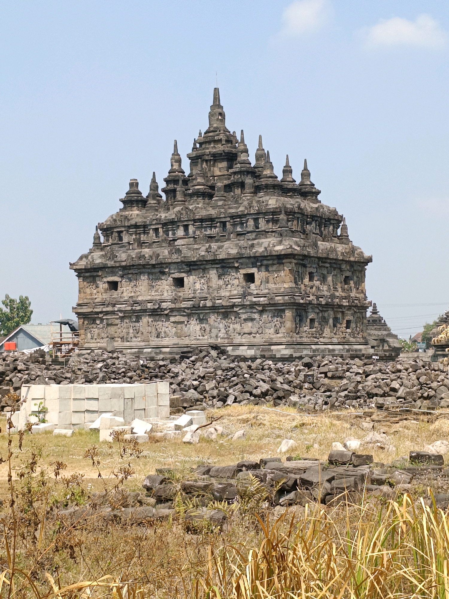 The image shows the Candi Plaosan temple complex in Bugisan, Indonesia. The temple is made of stone and has multiple tiers with intricate carvings. It is surrounded by piles of rubble and construction materials, suggesting ongoing restoration or archaeological work. The environment is dry and grassy, with a clear blue sky overhead. In the background, there are some trees and buildings, indicating the temple is located near a populated area. There are no people visible in the image.