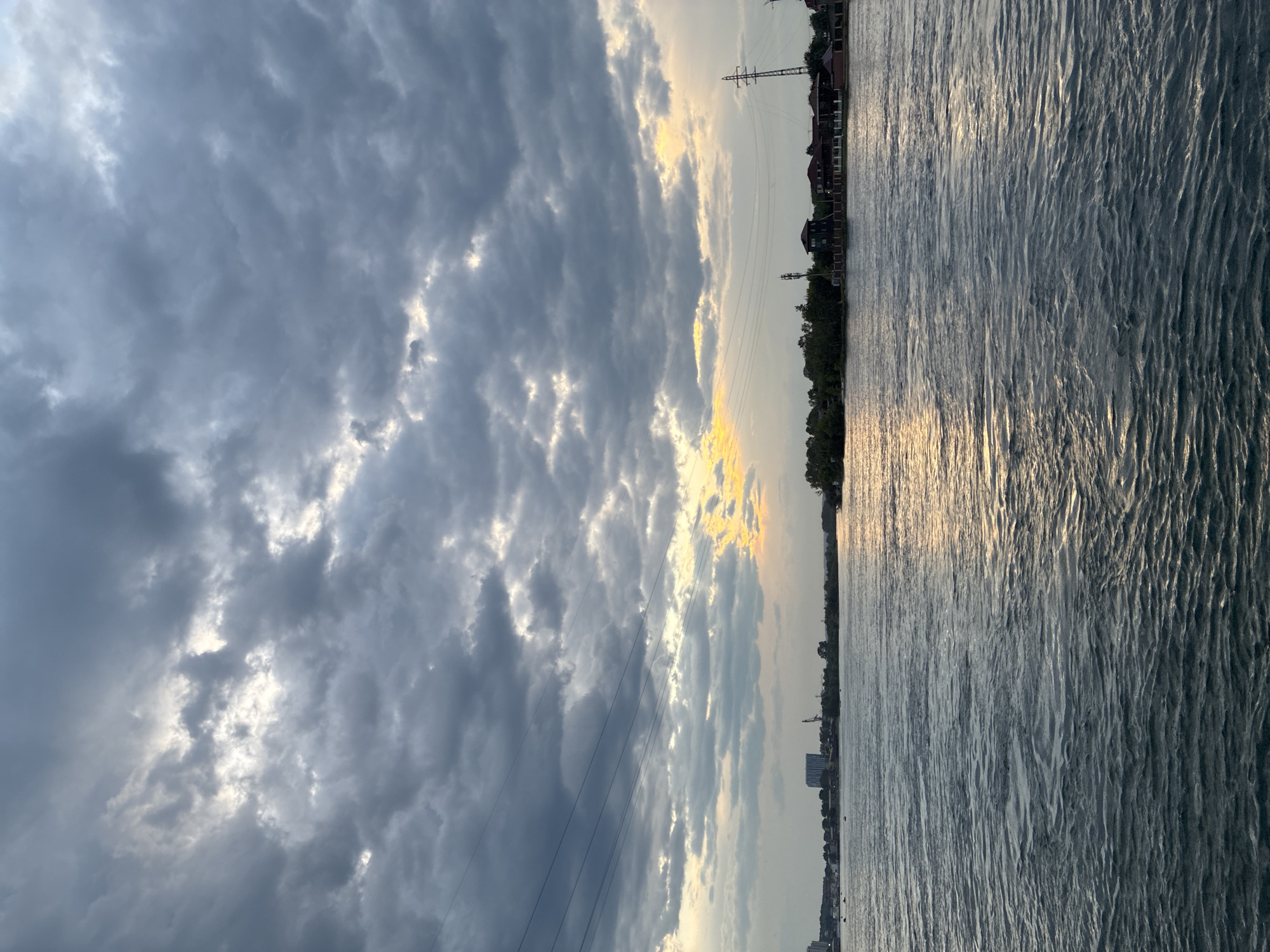 The image depicts a wide river flowing through an urban landscape, likely Irkutsk, Russia, at dusk. The sky is filled with dramatic, heavy grey clouds, with patches of sunlight breaking through, casting a warm glow on the water's surface. Power lines stretch across the upper portion of the frame, and a distant shoreline is visible, dotted with buildings, trees, and utility poles. The water is choppy, with small waves reflecting the subdued light. No people or specific actions are discernible in the scene.